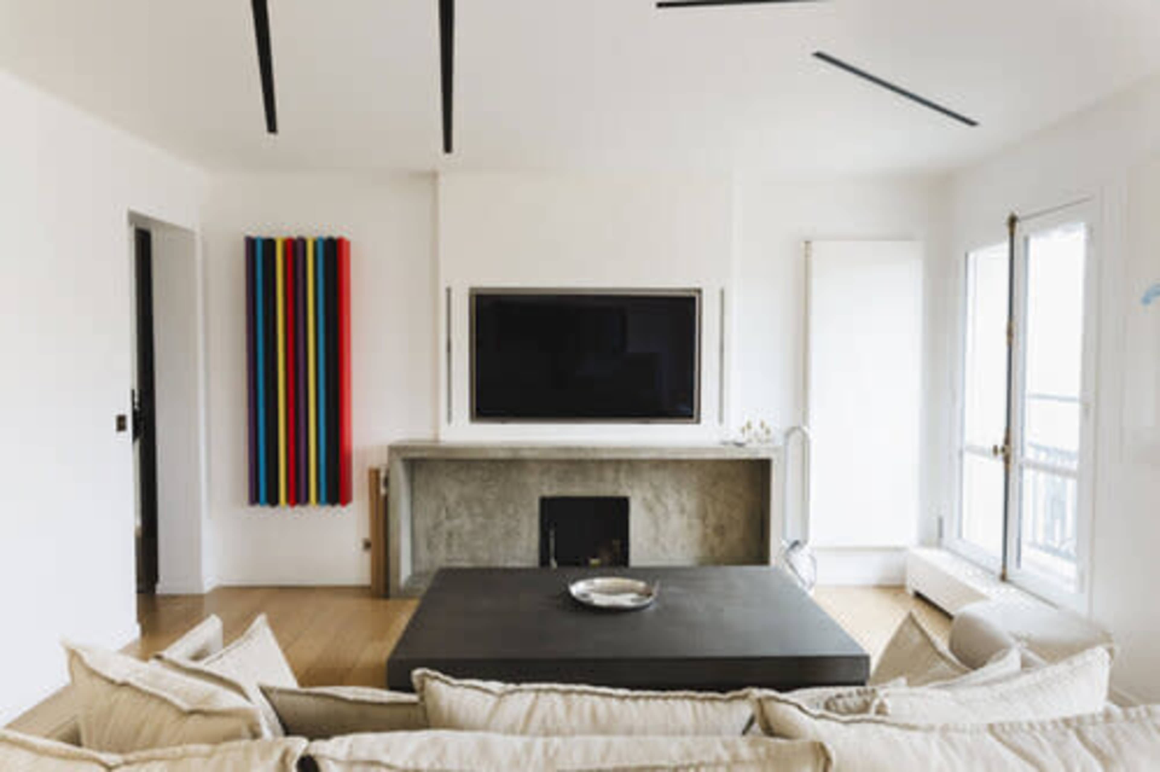 A modern living room features a large television mounted on a concrete wall, with a dark coffee table in front and colorful vertical art hanging nearby.