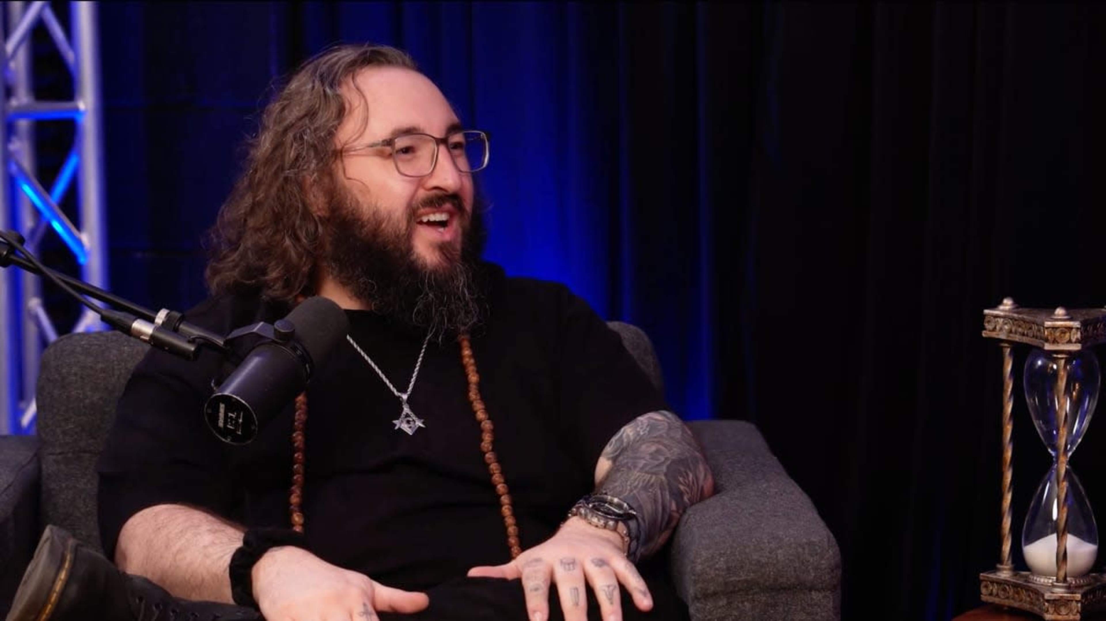 A man with long curly hair and glasses sits on a couch, speaking into a microphone in a dimly lit studio.