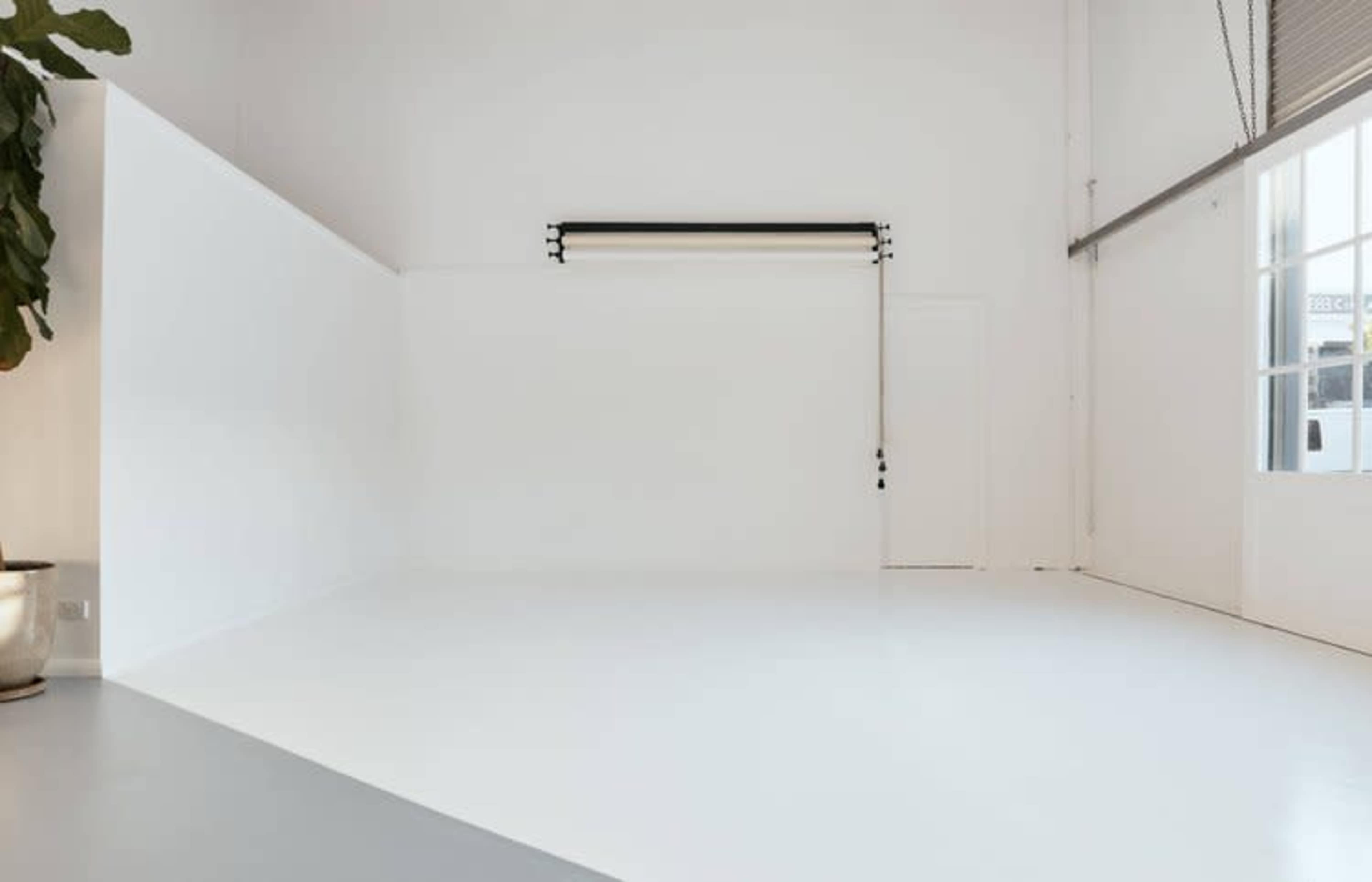 The image features a spacious studio with white walls and flooring, a curtain rod mounted on the wall, and large windows allowing natural light.