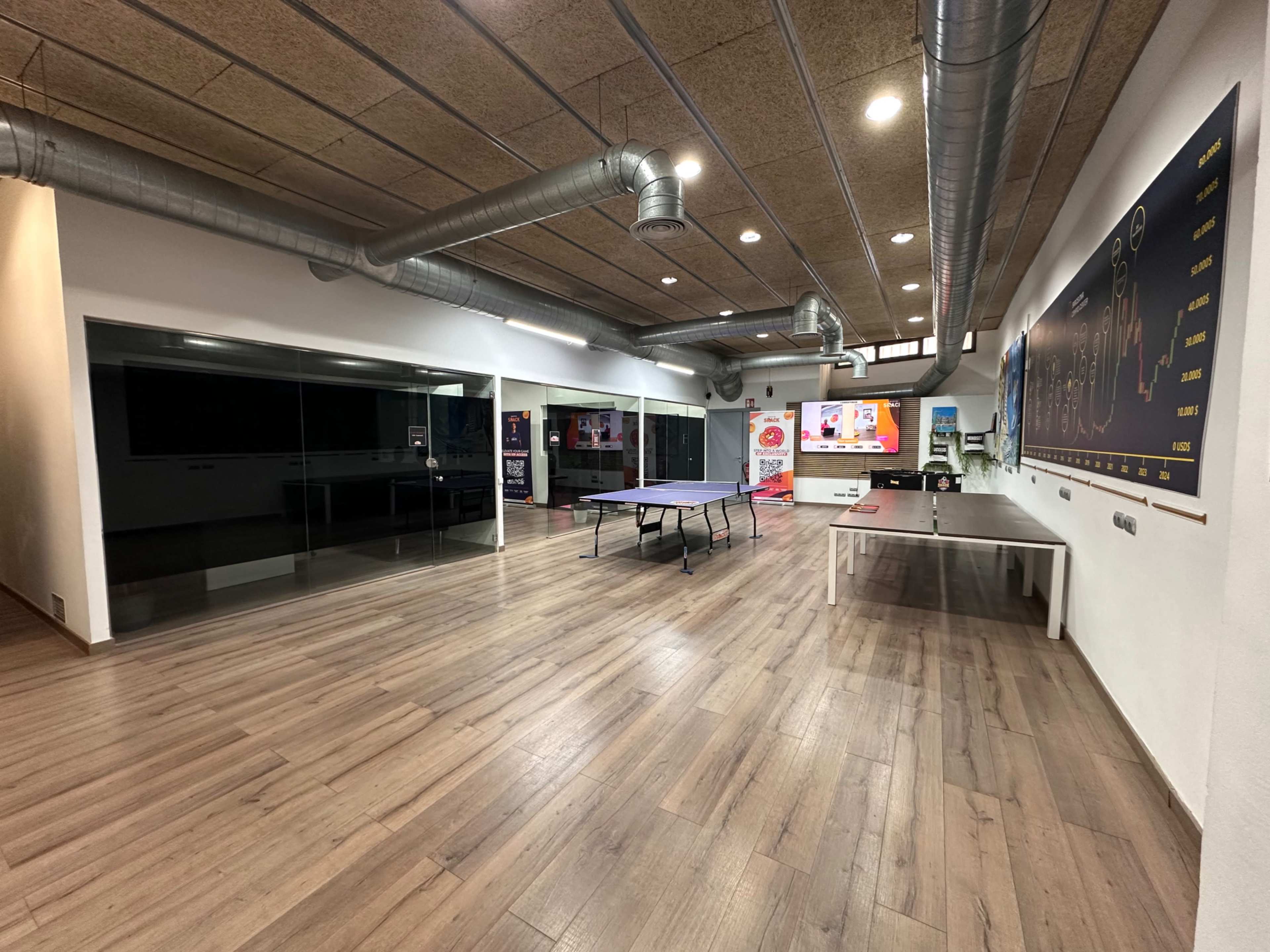 The image shows a spacious recreational room with wooden flooring, featuring a ping pong table, seating area, and large glass windows.