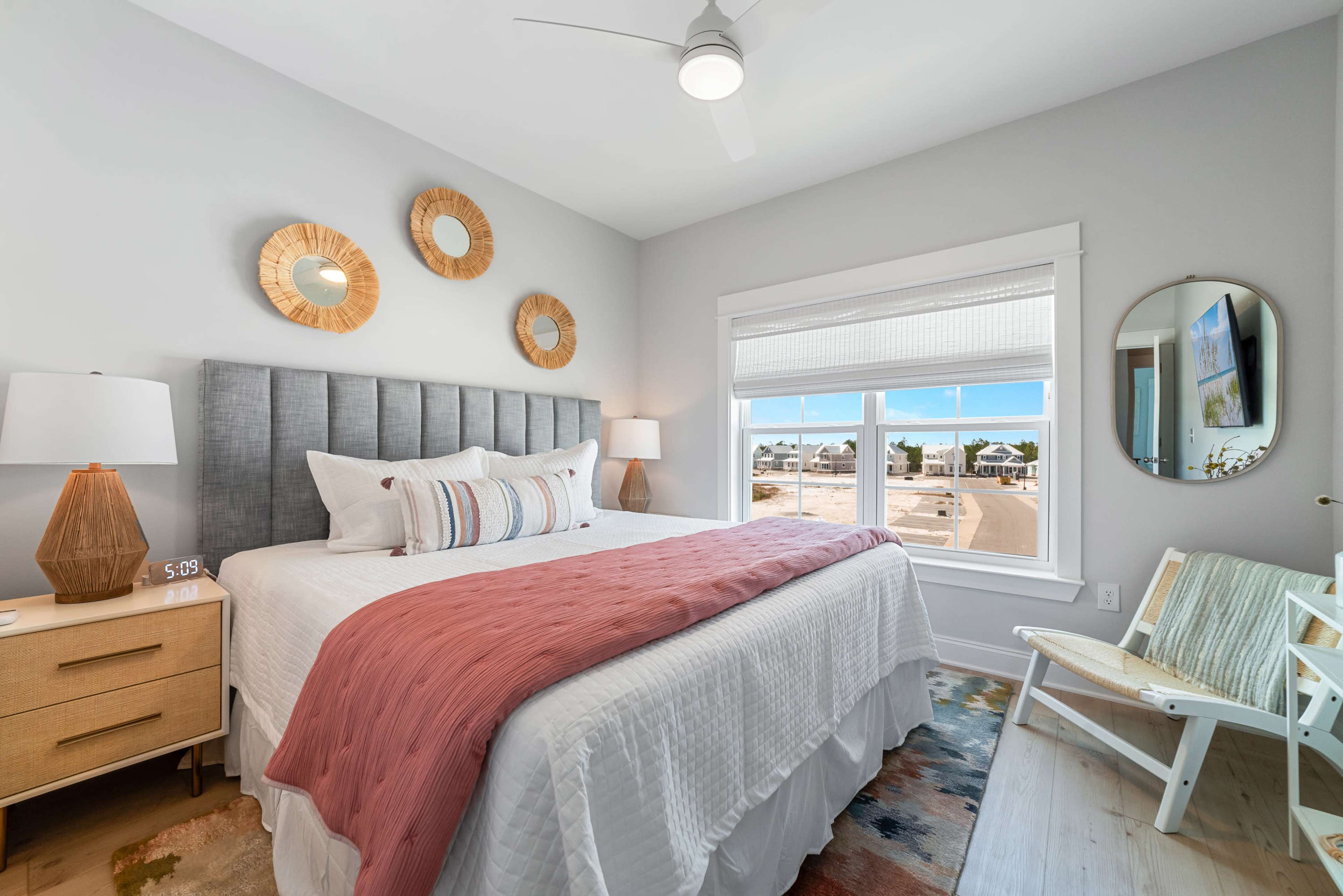 A neatly arranged bedroom features a large bed with a pink throw blanket, flanked by nightstands and lamps, alongside a window with three round mirrors on the wall.