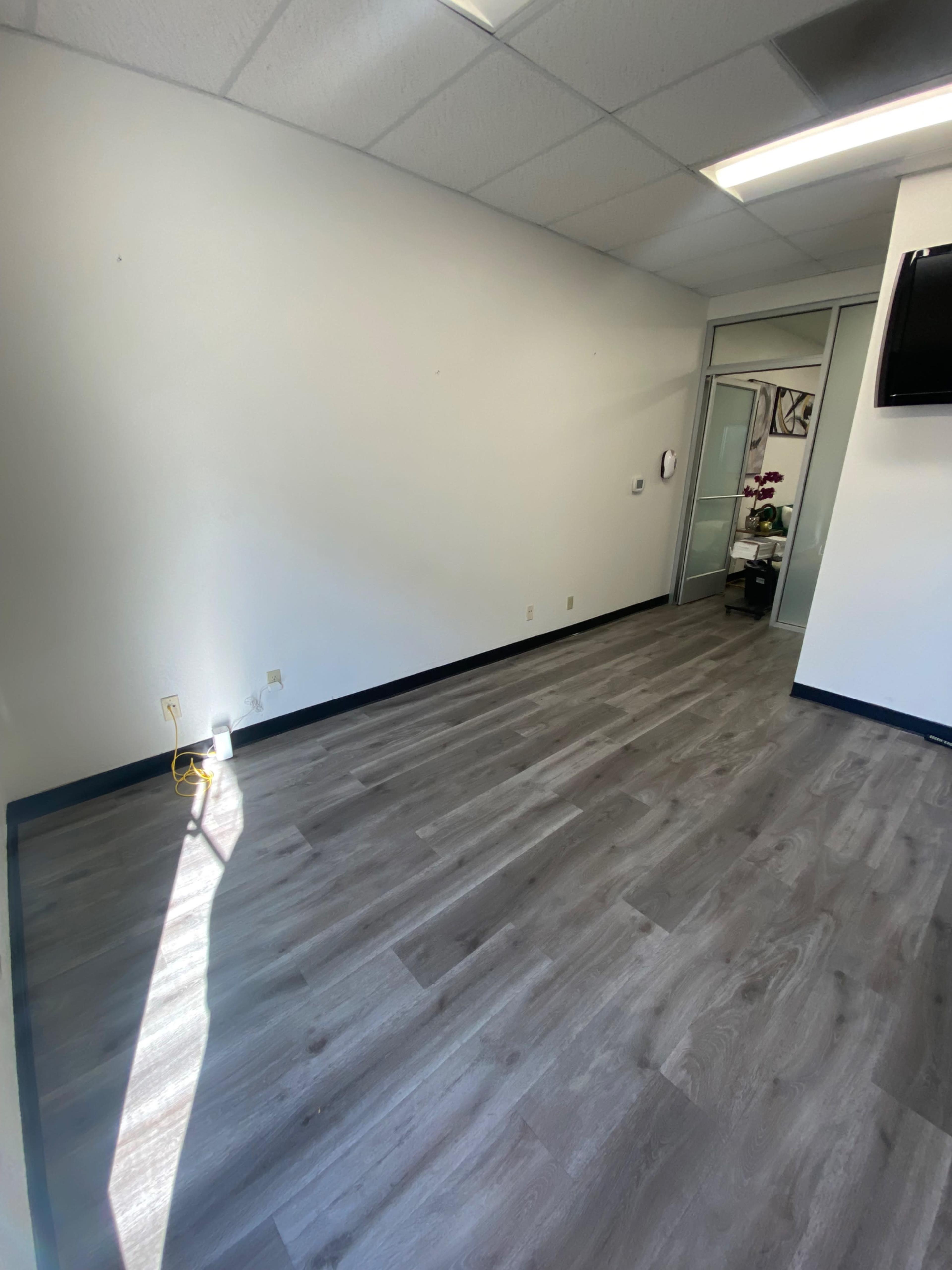 An empty office space with light-colored walls, wooden flooring, and a large window casting sunlight onto the floor.