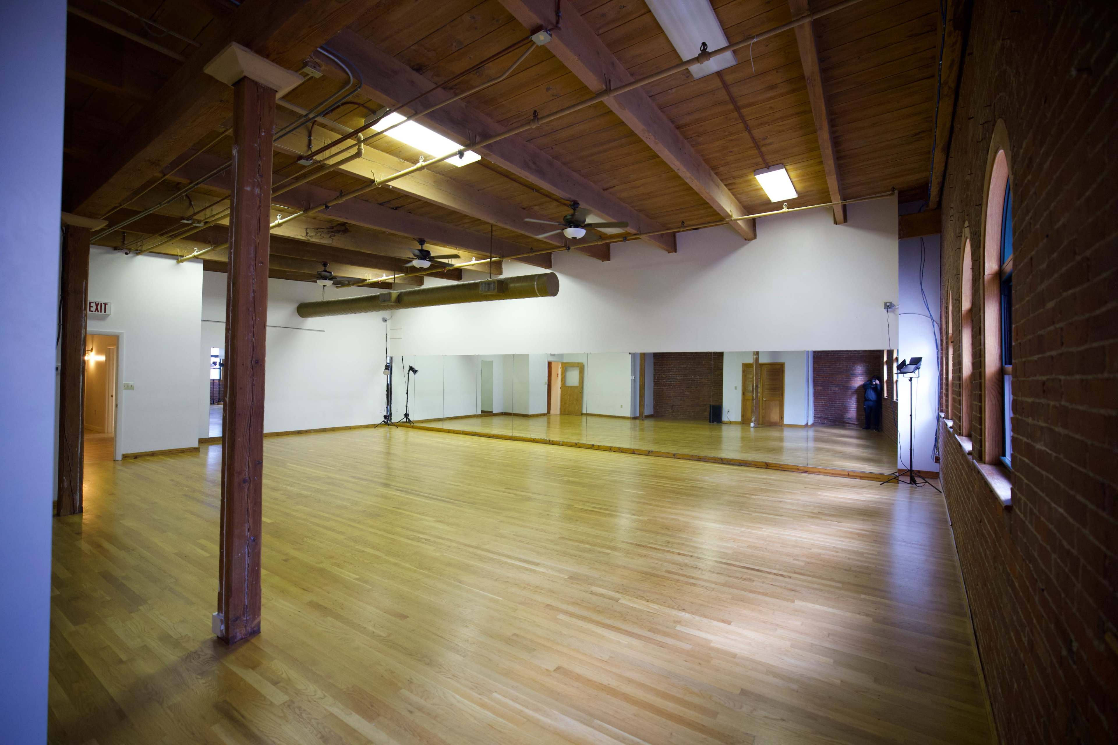 The image depicts an empty dance studio with wooden floors, exposed beams, and large mirrors on one wall.