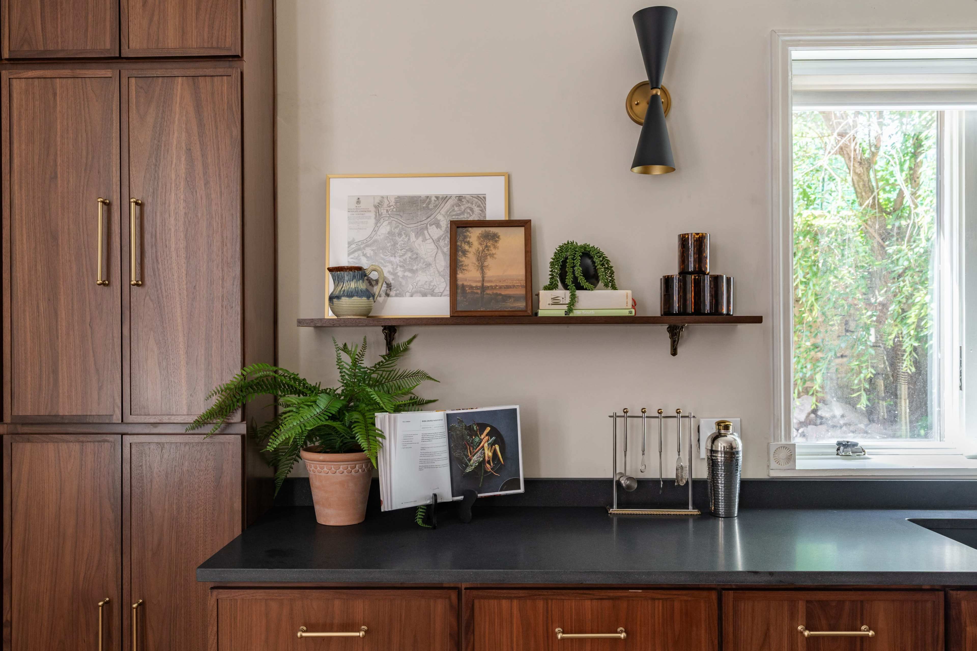The image shows a wooden kitchen cabinet with a black countertop, featuring a shelf with decorative items, a plant, and a nearby window with greenery outside.