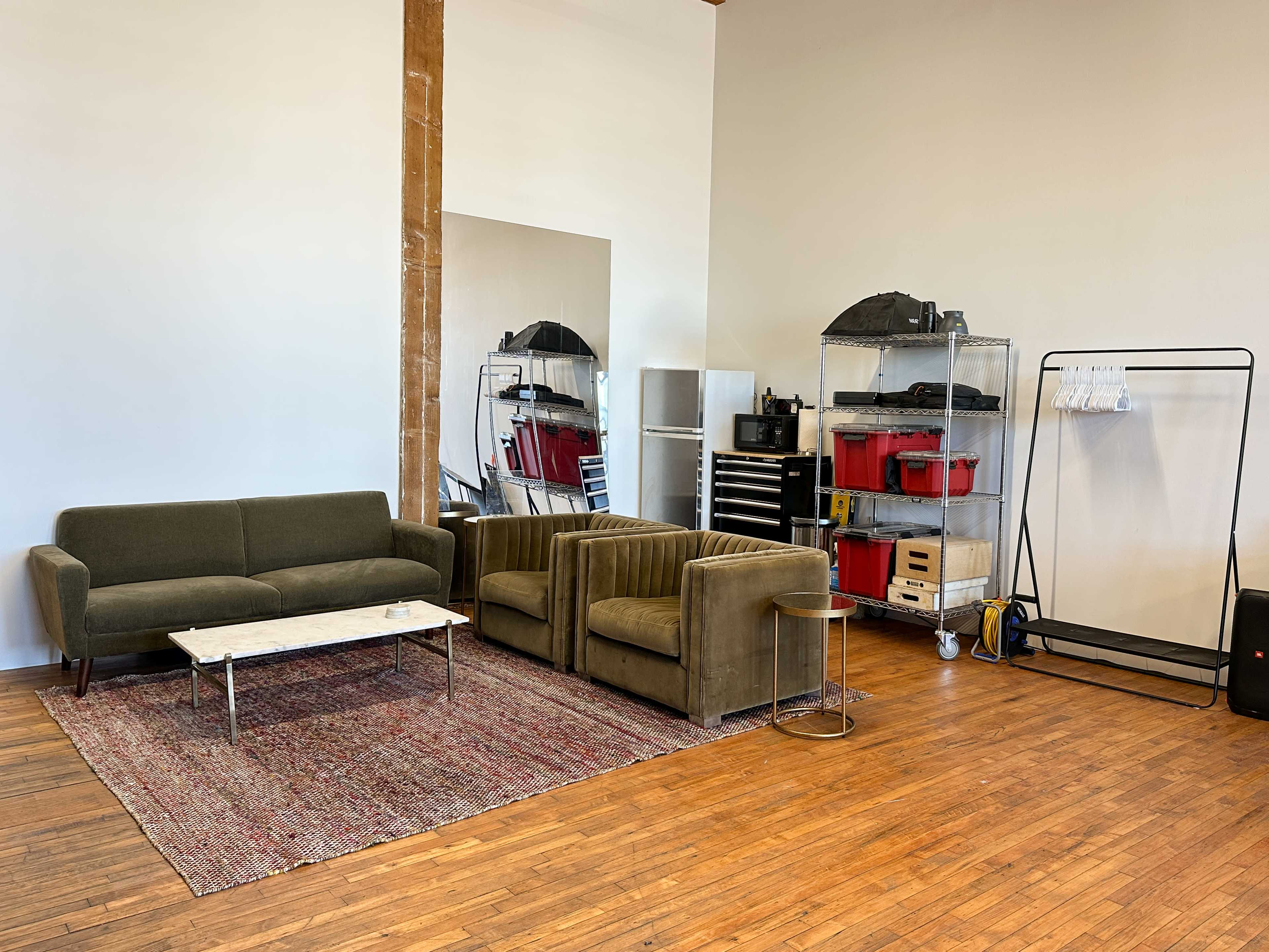 The image shows a minimalist room featuring a green sofa set, a coffee table on an area rug, a mirrored wall, and storage shelves filled with tools and equipment.