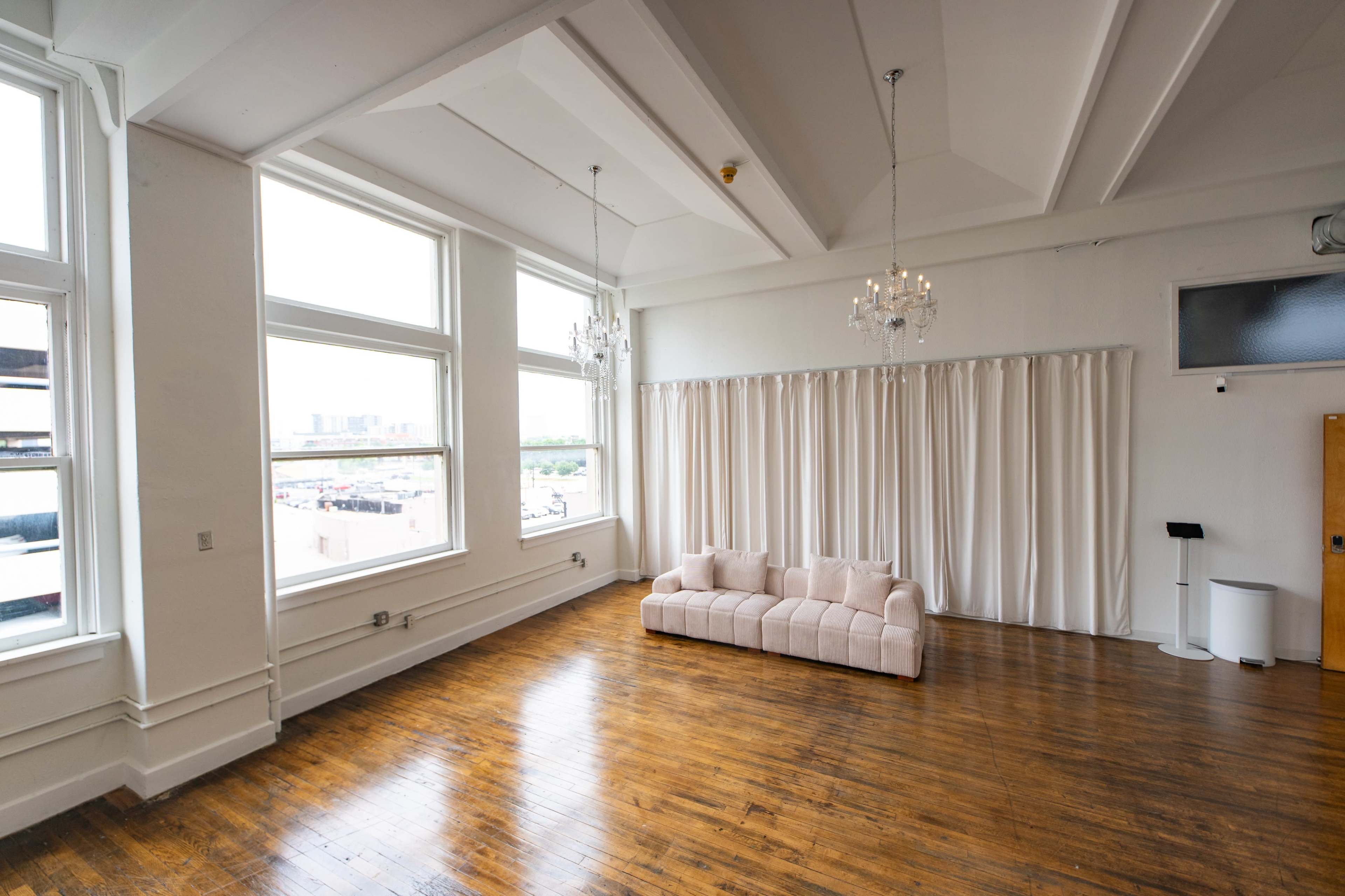 A spacious room features large windows, a light-colored sofa, and decorative chandeliers against a backdrop of white curtains and hardwood flooring.