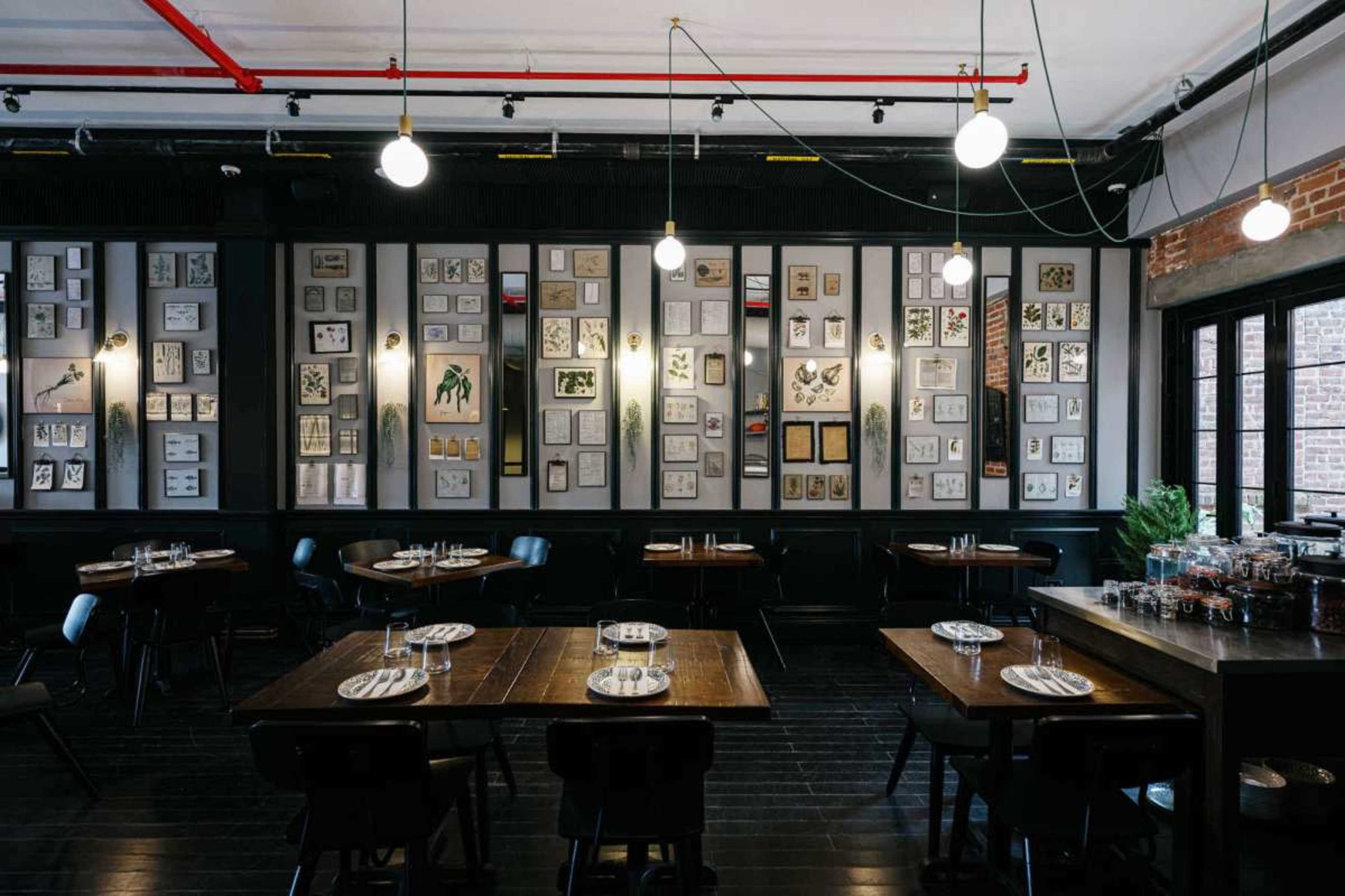The image shows the interior of a restaurant featuring wooden tables, black chairs, and a wall adorned with framed artworks and photographs.
