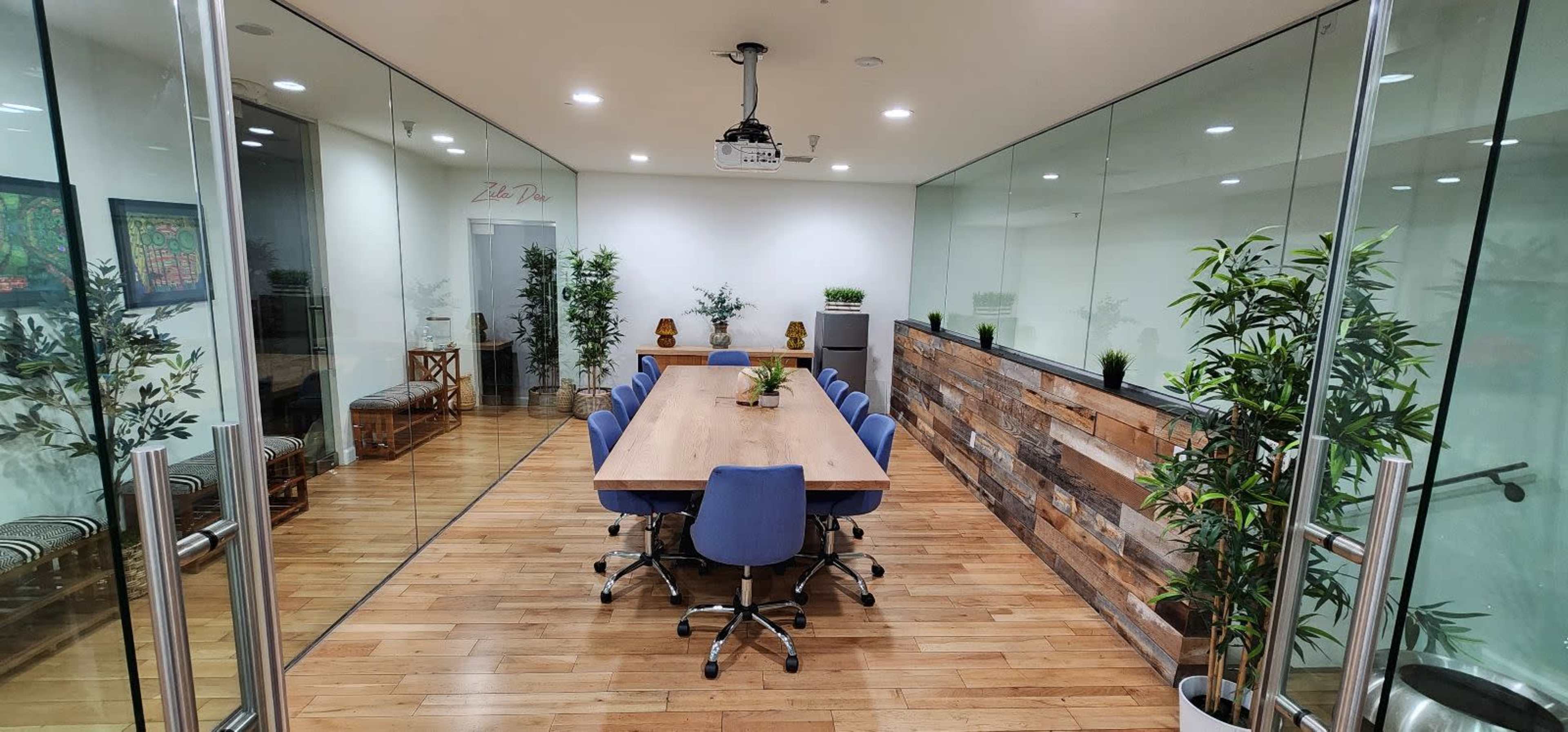 A modern conference room with a long wooden table surrounded by blue chairs and large glass walls, featuring decorative plants and a projector on the ceiling.