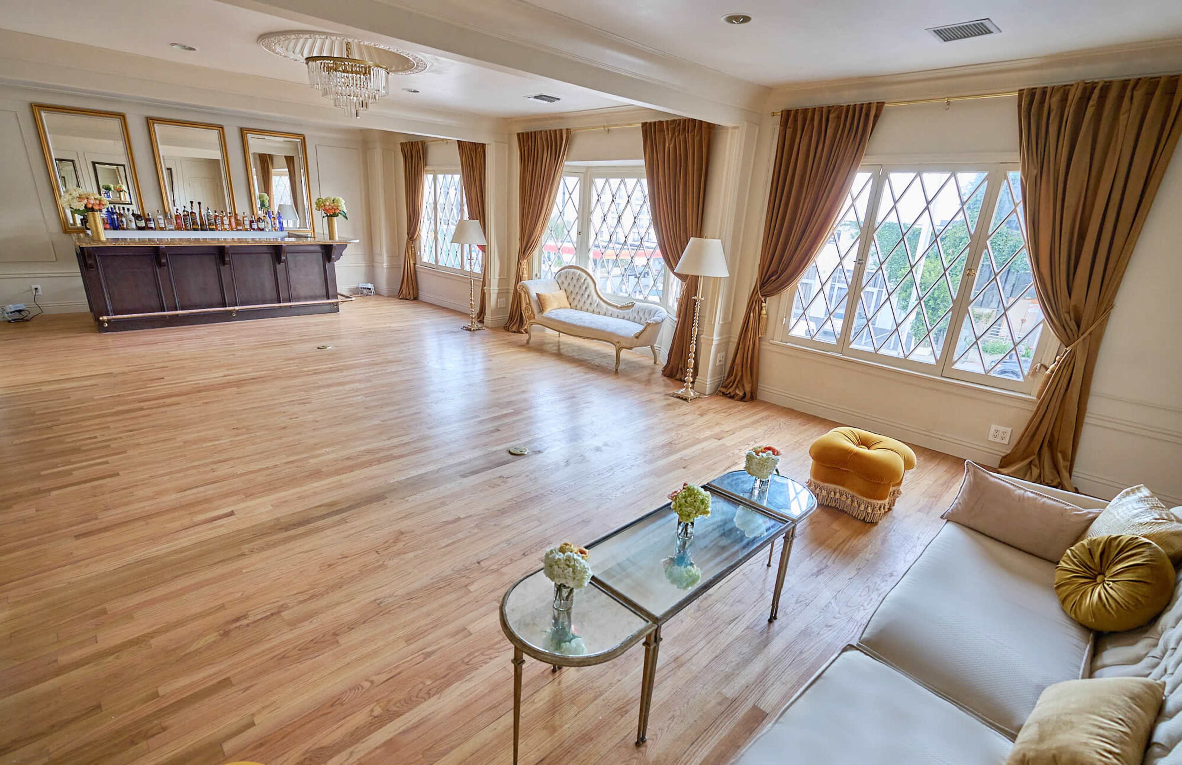 The image shows a spacious room with hardwood flooring, large windows adorned with drapes, a bar at the back, and a seating area that includes a couch and a yellow ottoman.