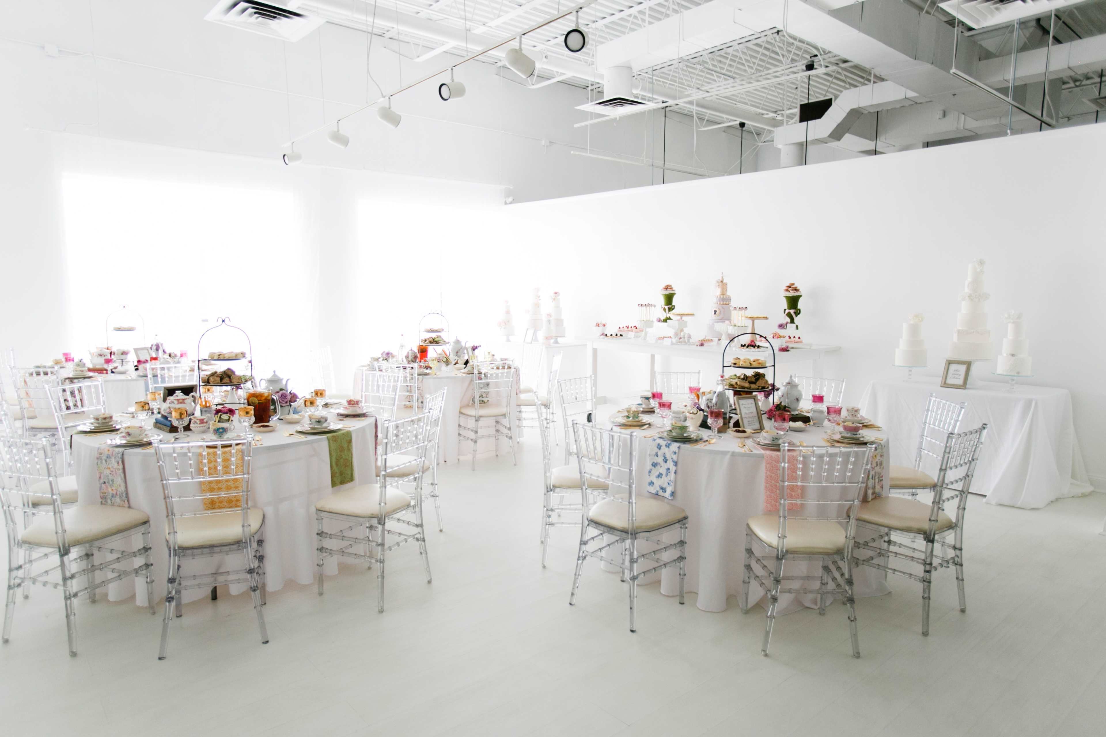 A bright, modern event space set up for a gathering, featuring neatly arranged round tables with white tablecloths, chairs, and a variety of food displays.