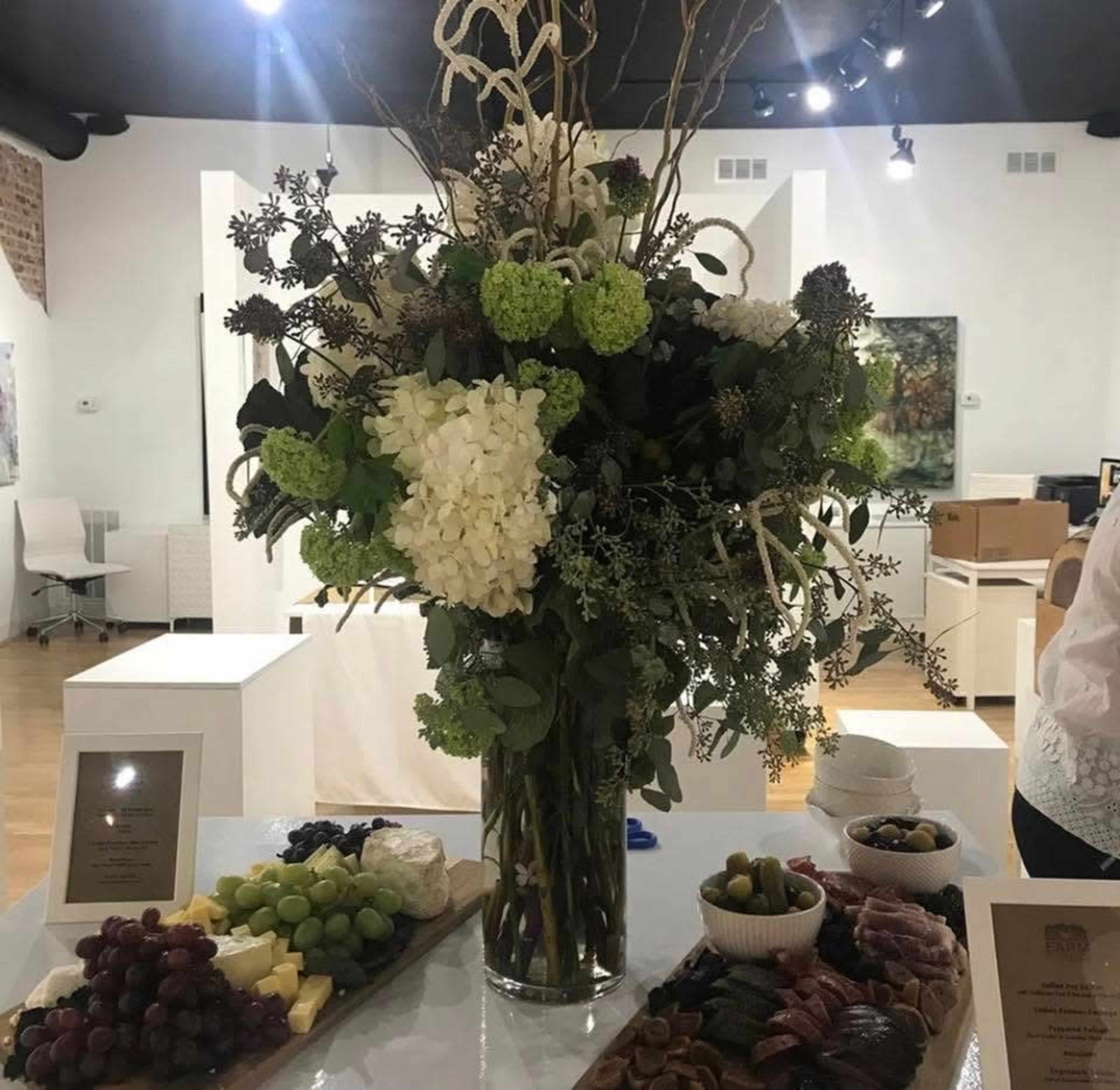 A large floral arrangement stands in a glass vase on a table with an assortment of cheeses, fruits, and olives arranged on a wooden platter.