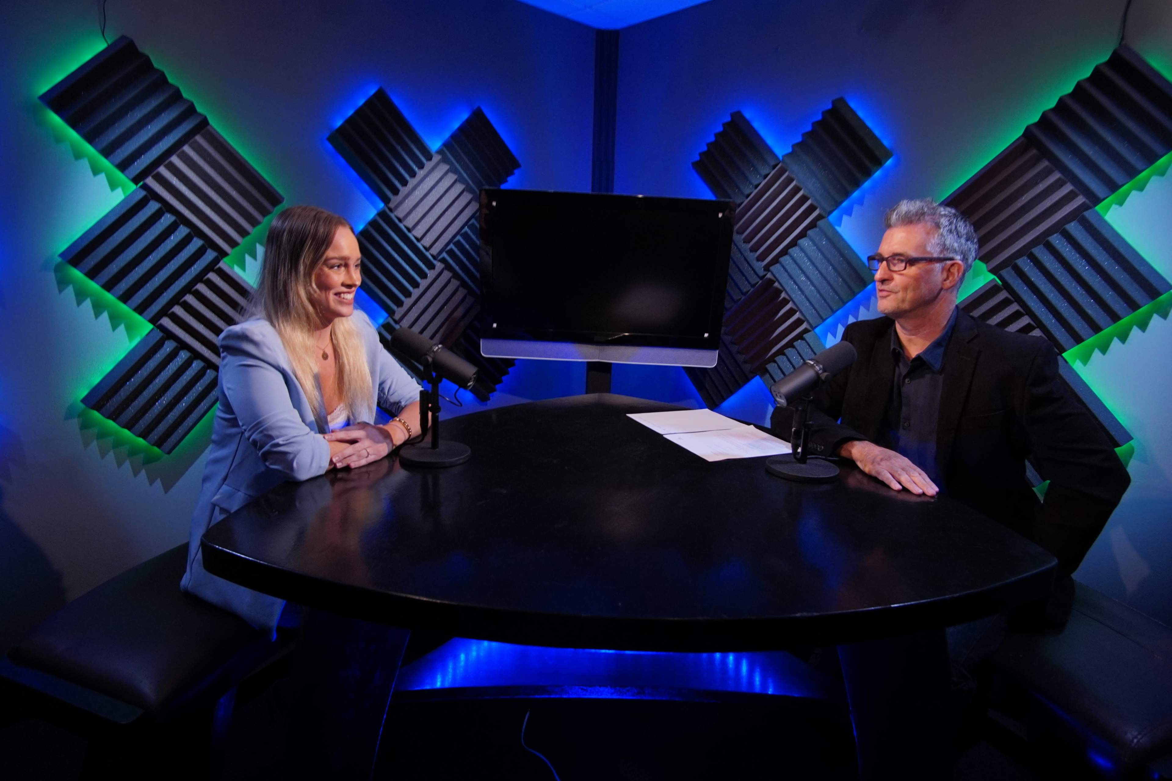 A man and a woman sit at a table with microphones in a recording studio featuring colorful LED lighting and soundproofing panels.