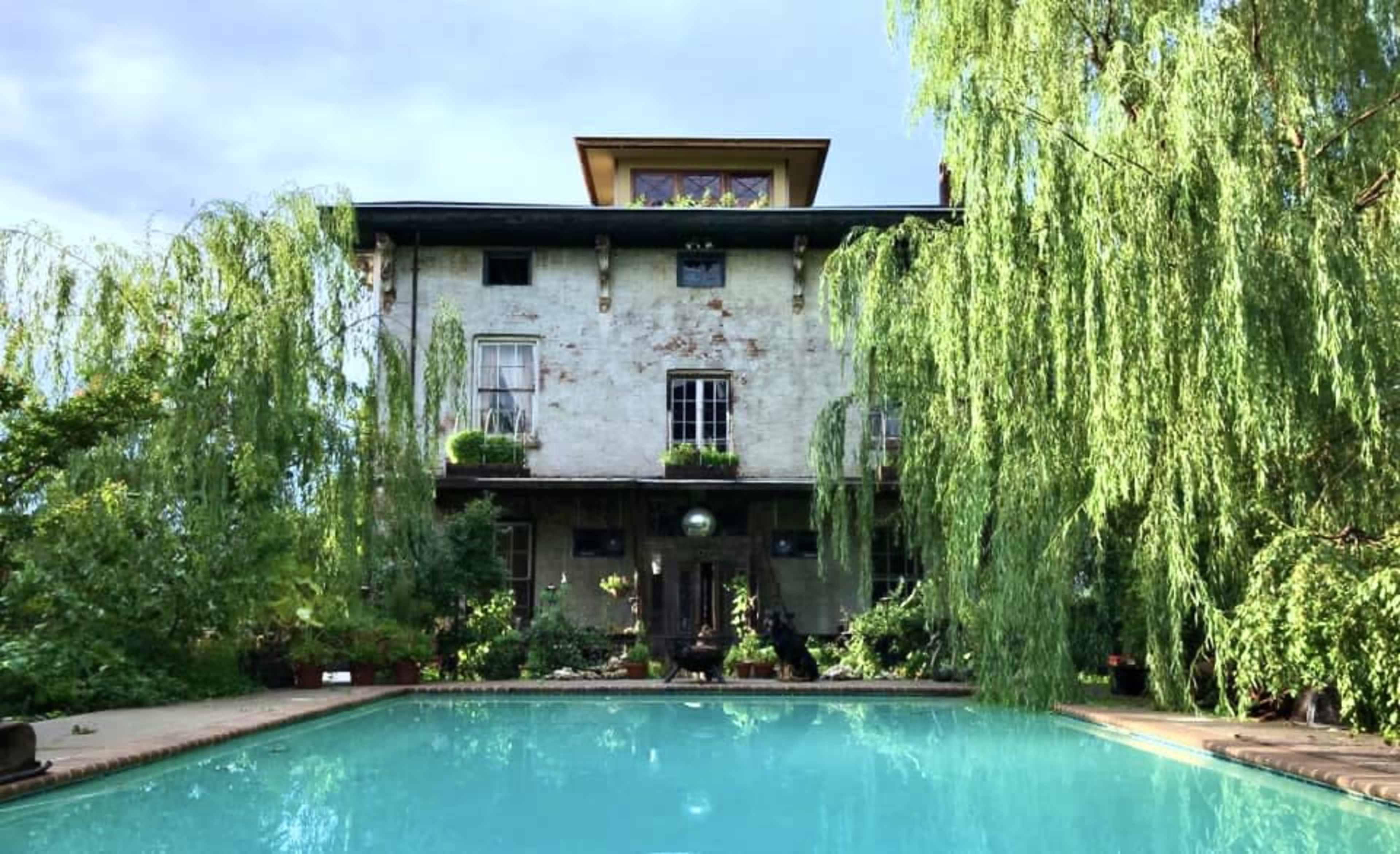 A historic brick building with tall windows is situated behind a turquoise swimming pool and surrounded by weeping willow trees.