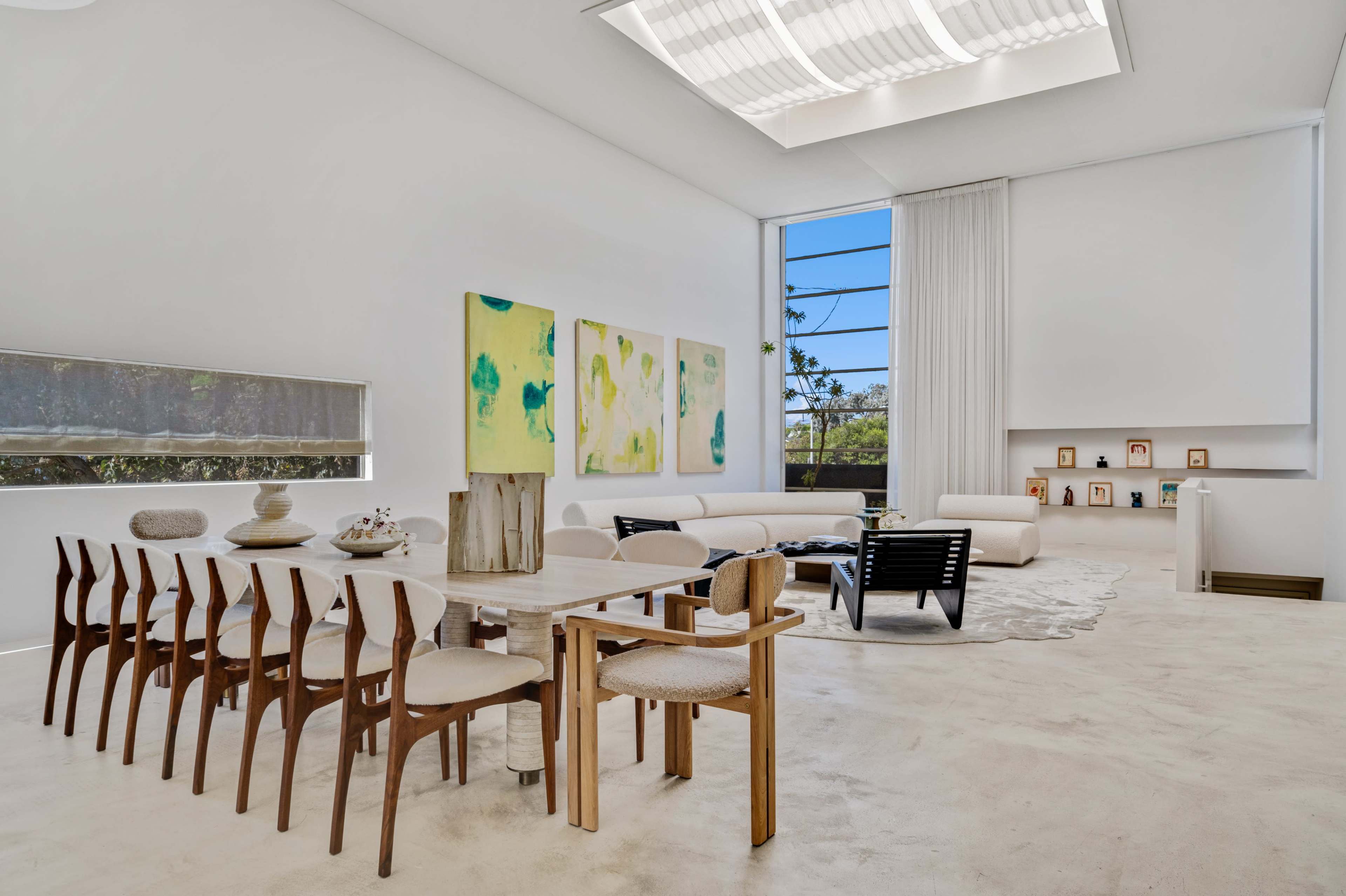 A spacious, minimalist living area features a dining table with chairs, large windows allowing natural light, and abstract paintings on the wall.