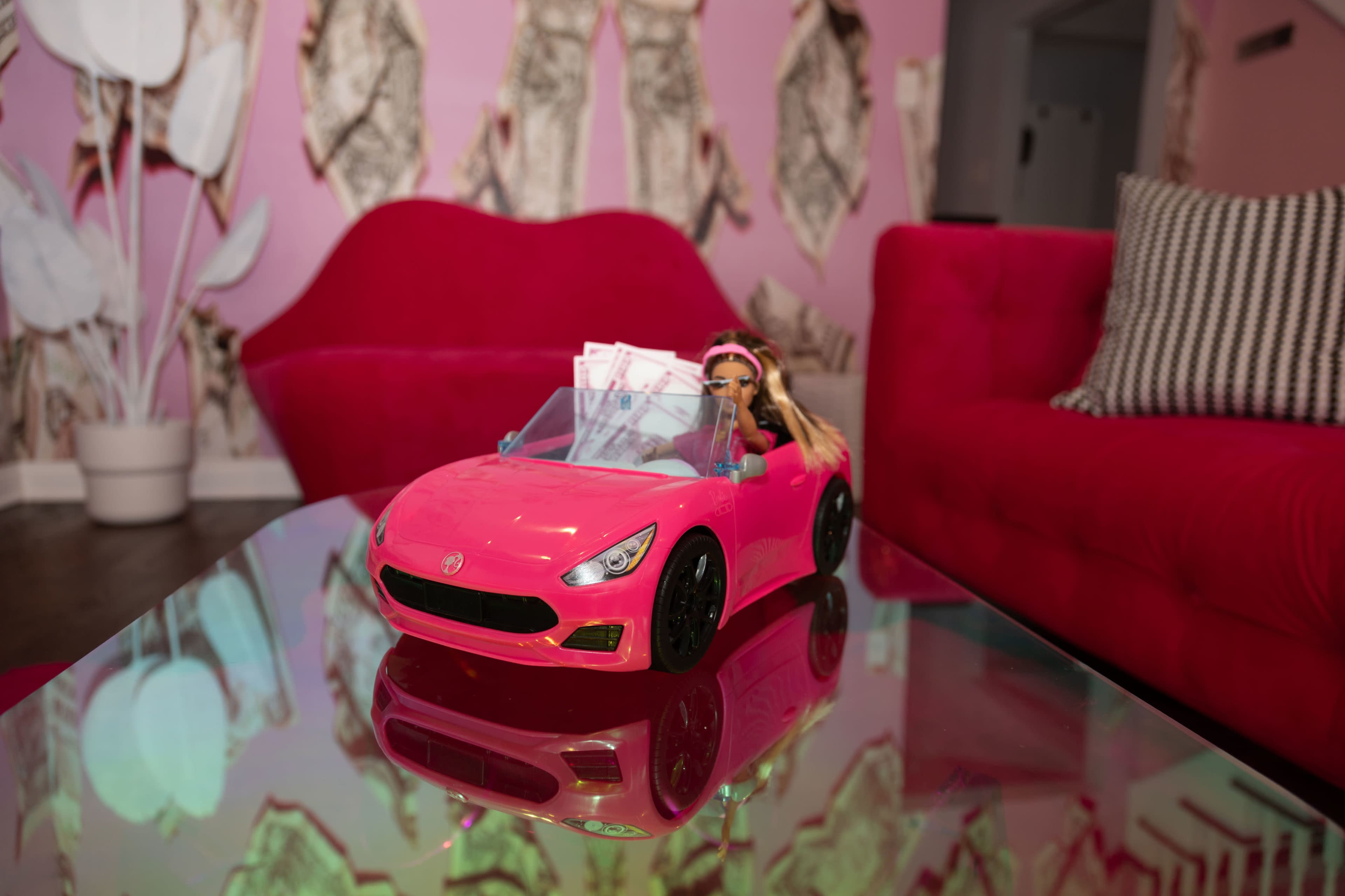 A pink toy convertible car with a doll inside is parked on a reflective glass coffee table in a room featuring vibrant pink decor.