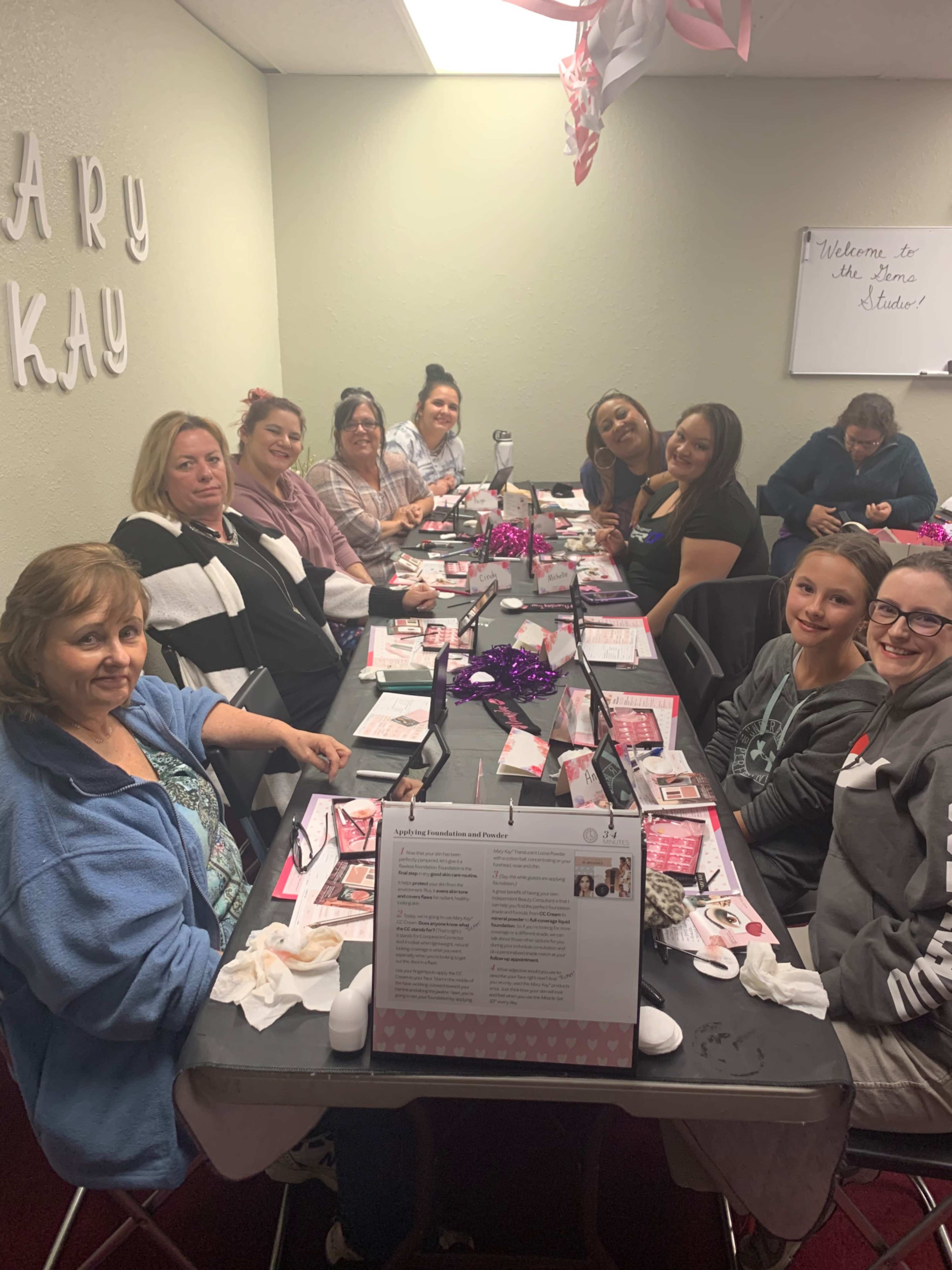 A group of eleven women sits around a table decorated with pink accents, engaged in activities and conversations during a gathering.