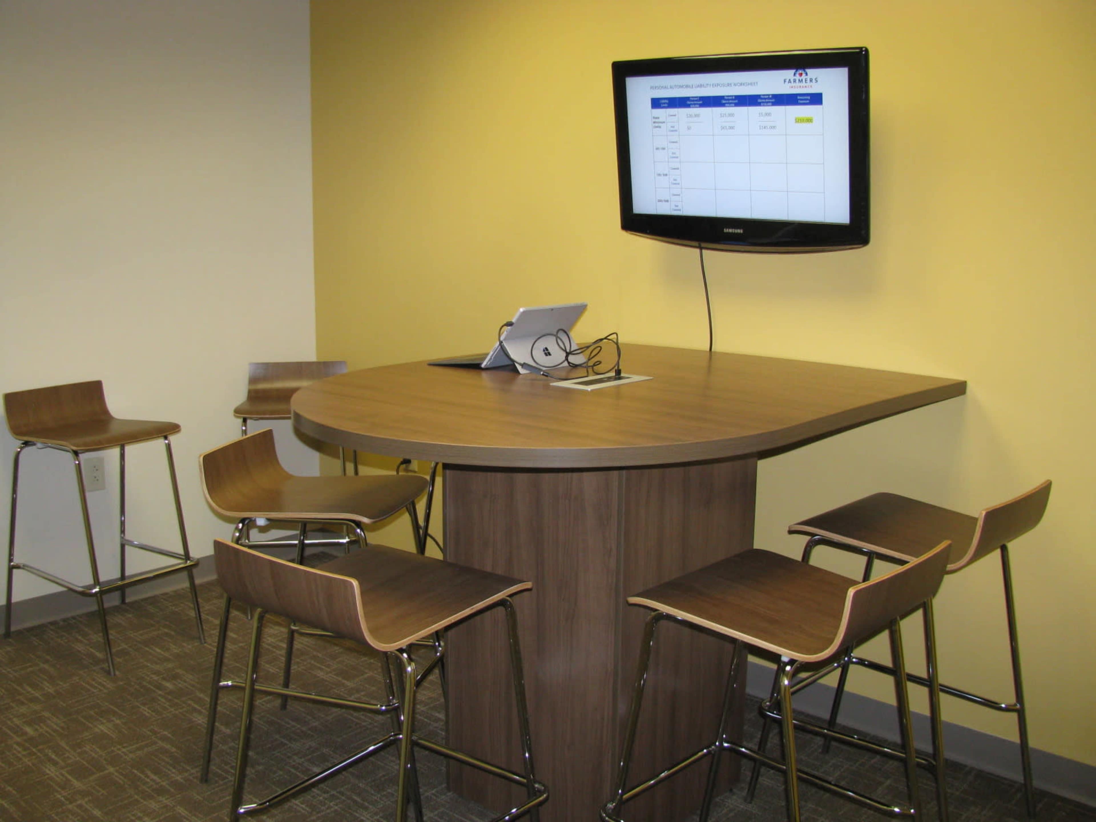 A modern conference area features a brown table with high stools and a wall-mounted television displaying a spreadsheet.