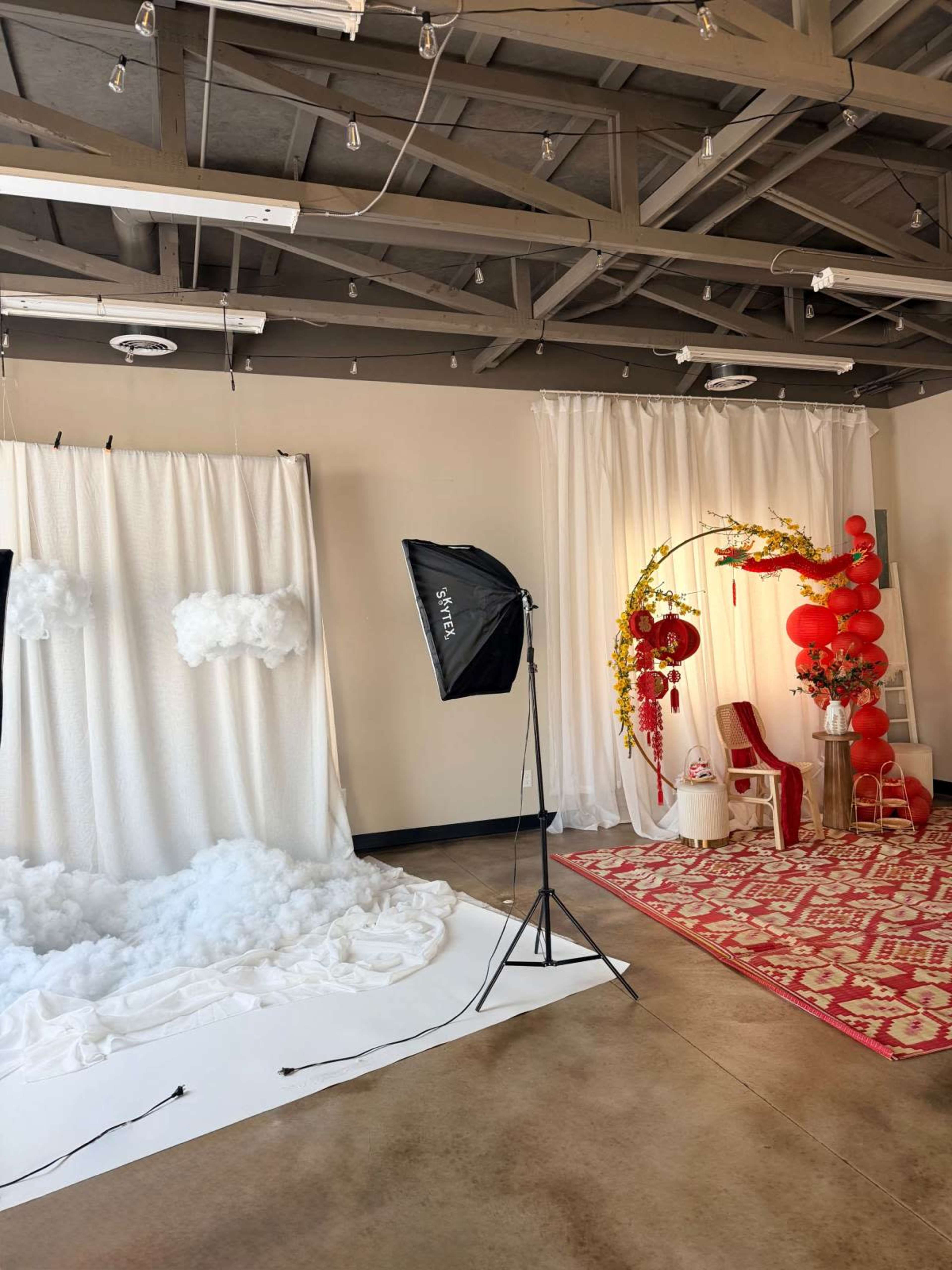 The image shows a photography studio with a backdrop featuring clouds, a lighting umbrella, and a decorated corner with red balloons and a chair.