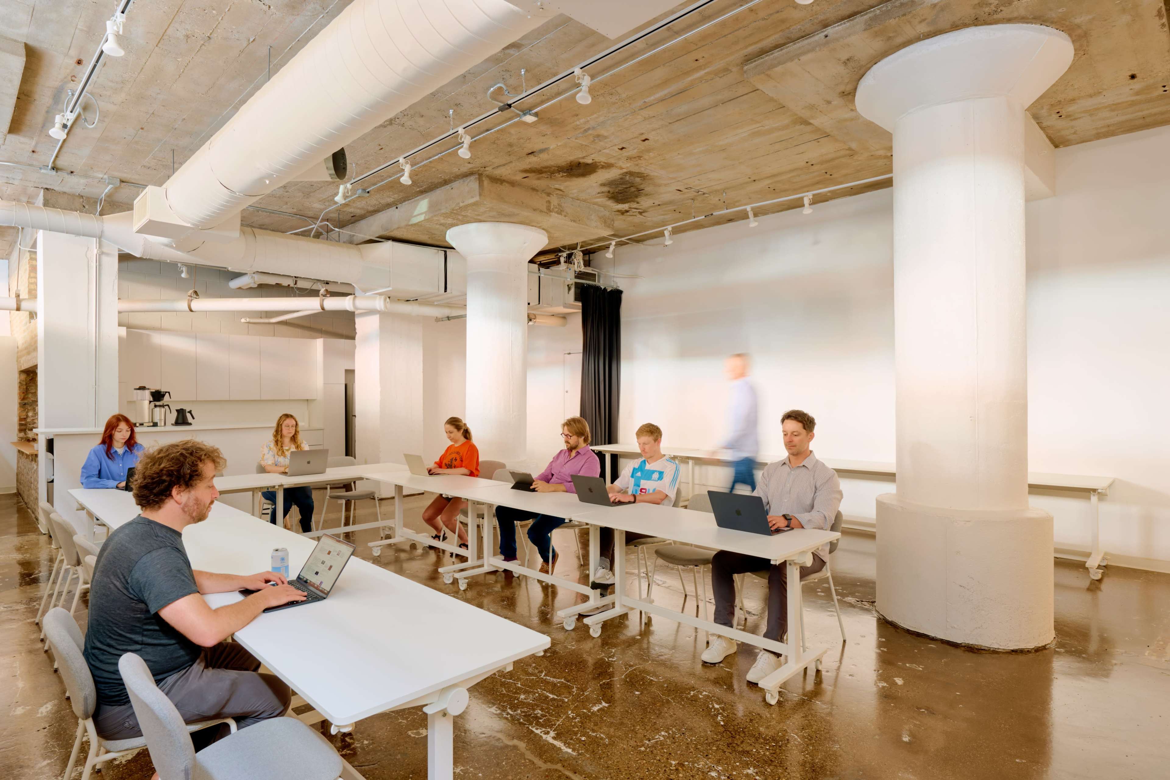 A group of six individuals is seated at long tables in a spacious, modern meeting room with concrete pillars and a polished floor, while one person walks in the background.