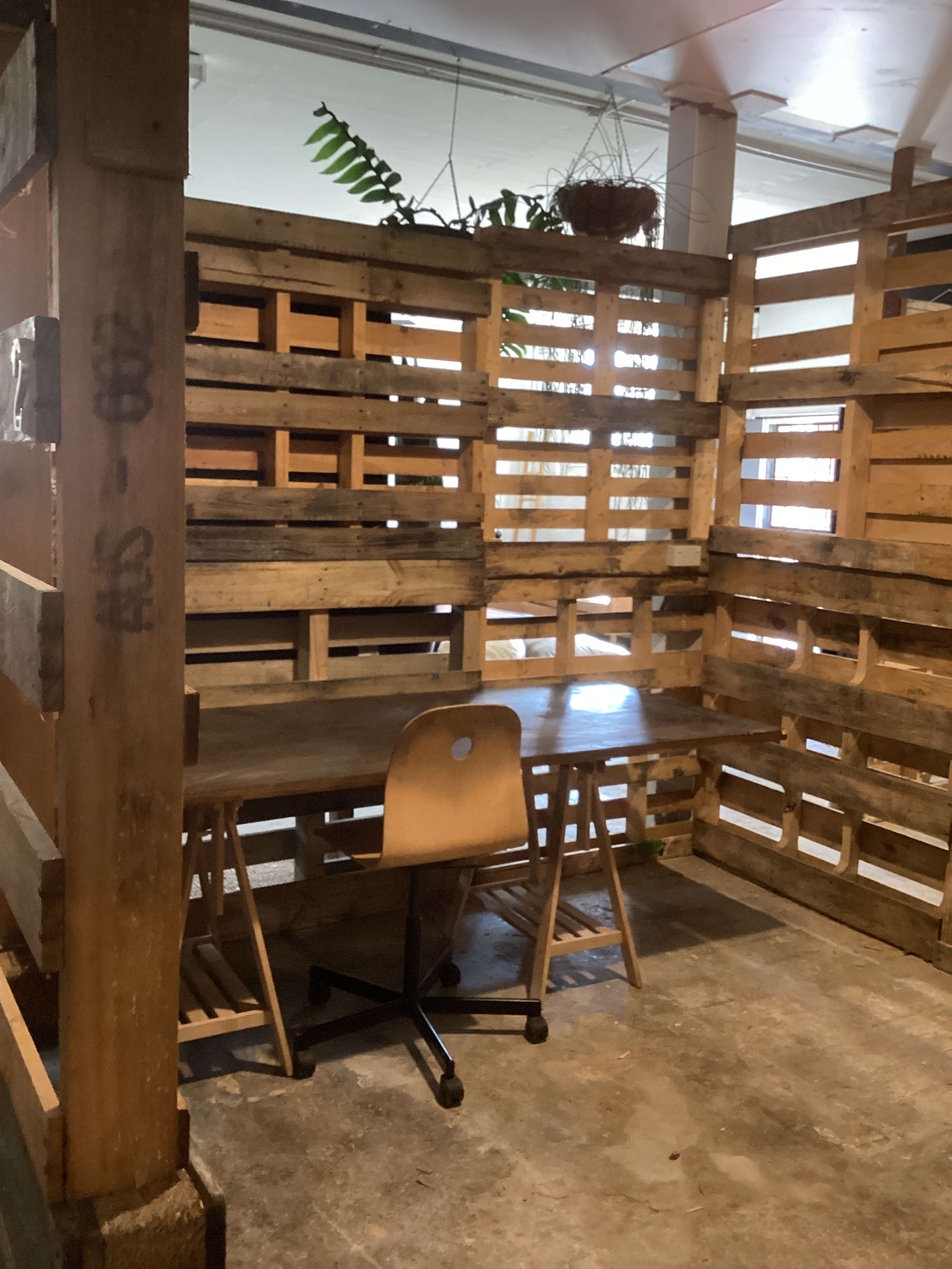 The image shows a small workspace enclosed by wooden pallets, featuring a table and a chair.
