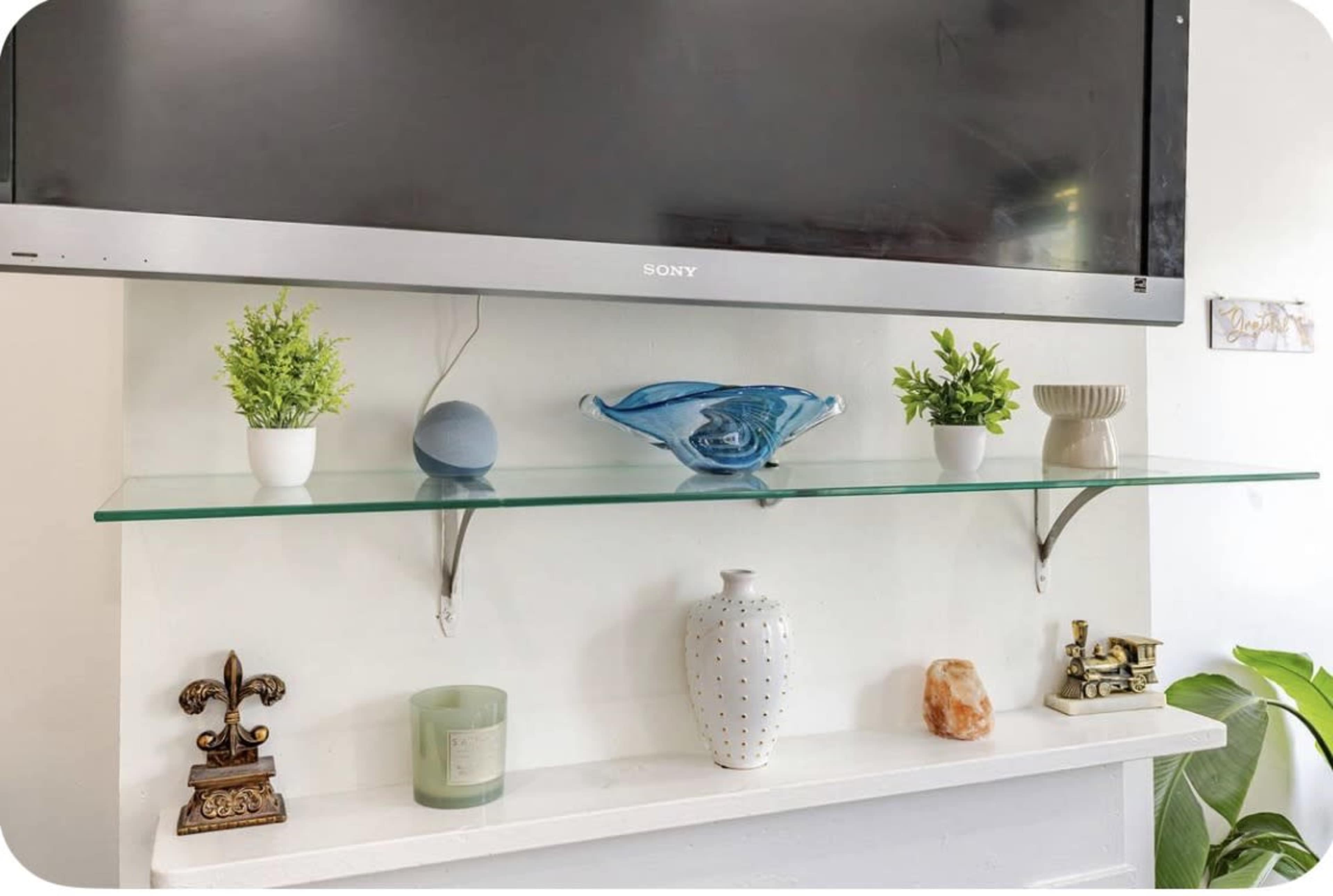 A glass shelf displays several decorative items, including potted plants, a blue glass bowl, a vase, a candle, and a small statue, beneath a mounted television.