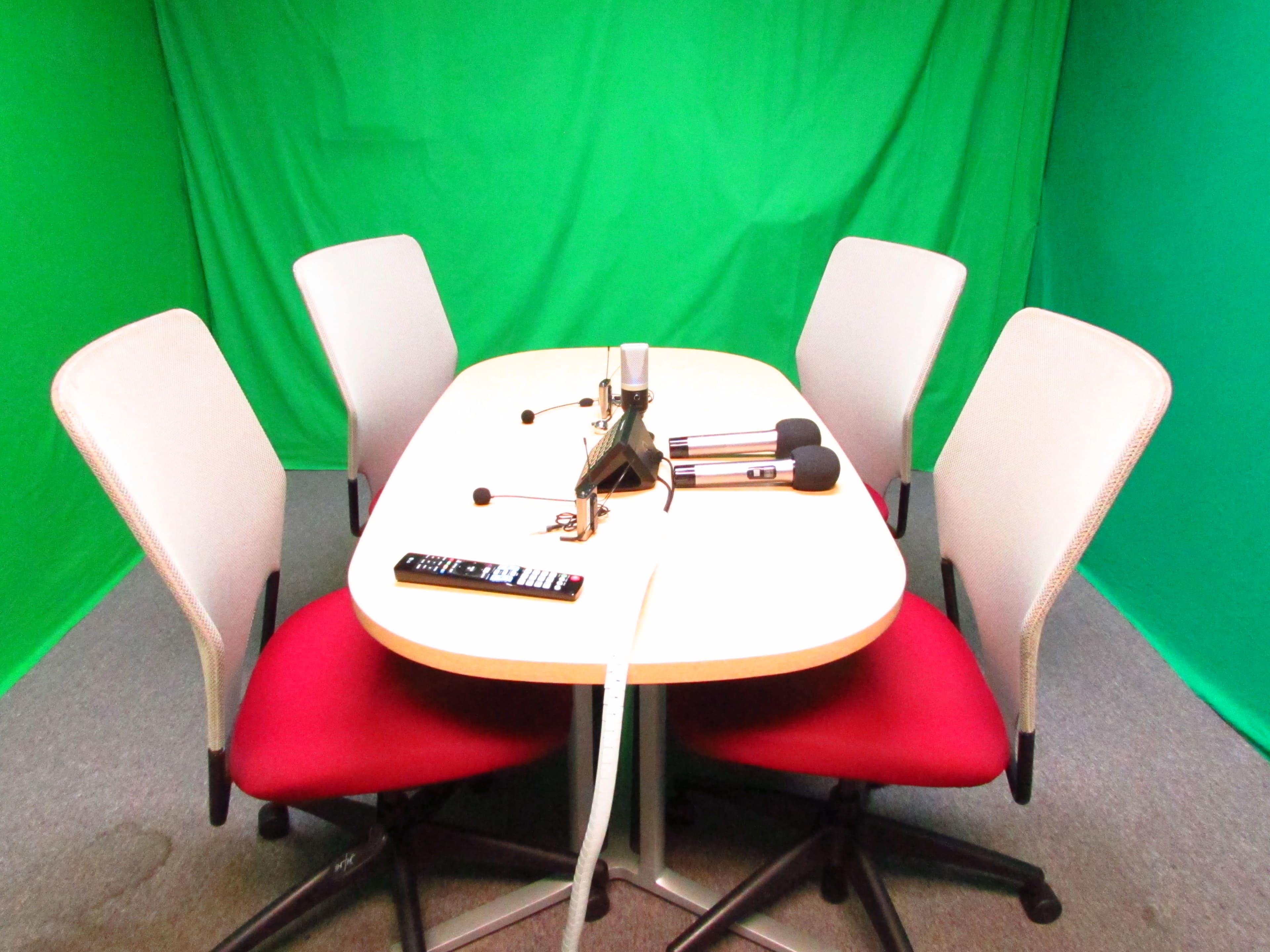 A table with four chairs is set up against a green backdrop, with microphones and a remote control arranged on the table.