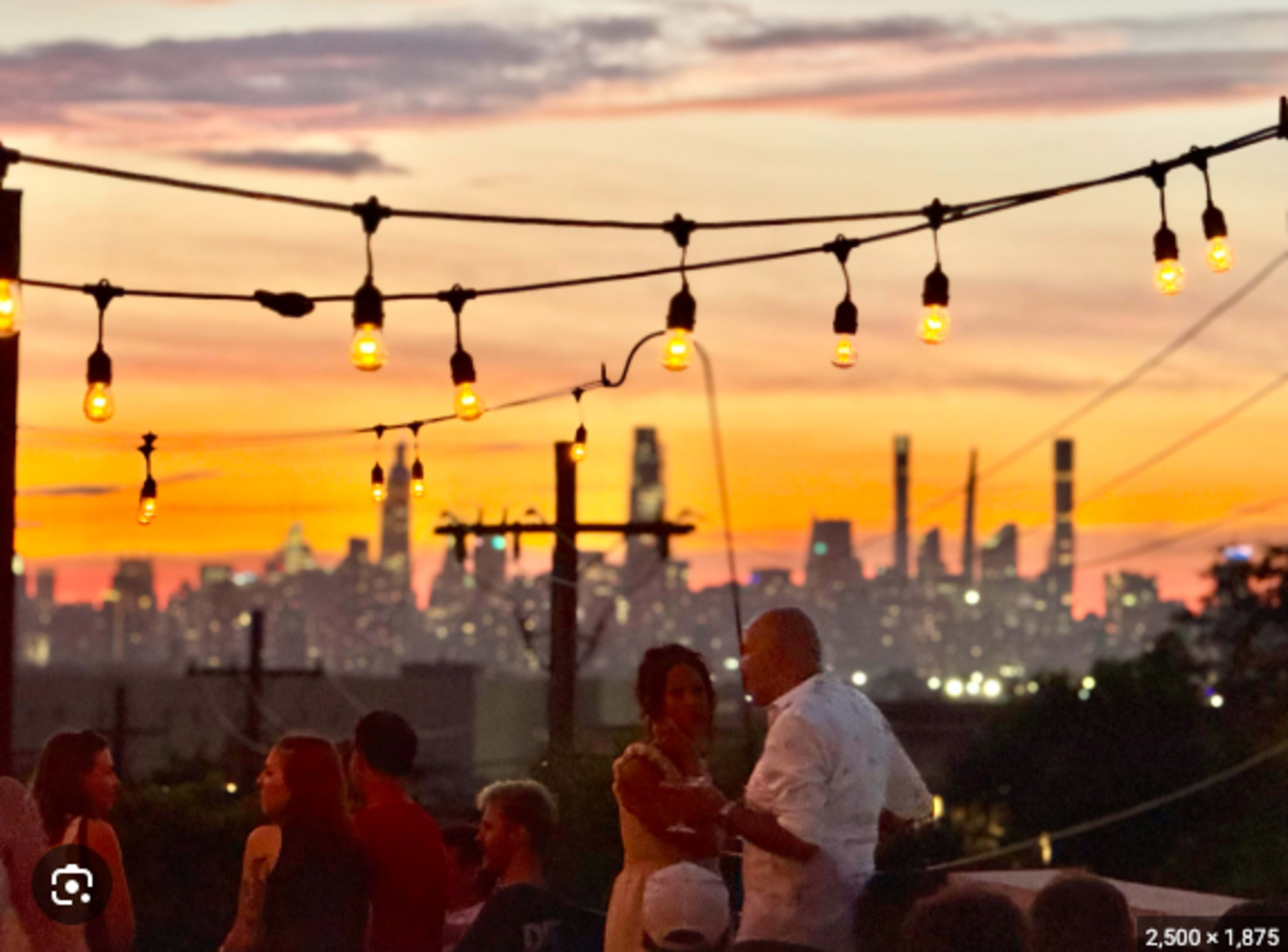 People gather at an outdoor event during sunset, with a city skyline illuminated in the background and string lights overhead.