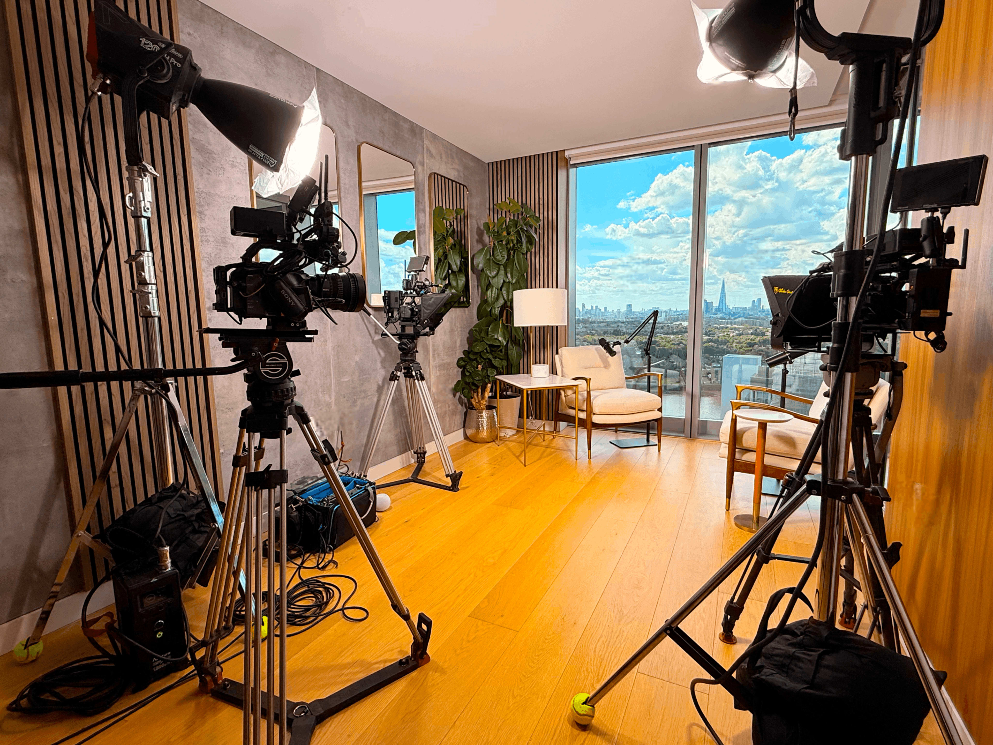 The image shows a modern studio setup with multiple cameras and lighting equipment aimed at a seating area with a view of a city skyline through large windows.