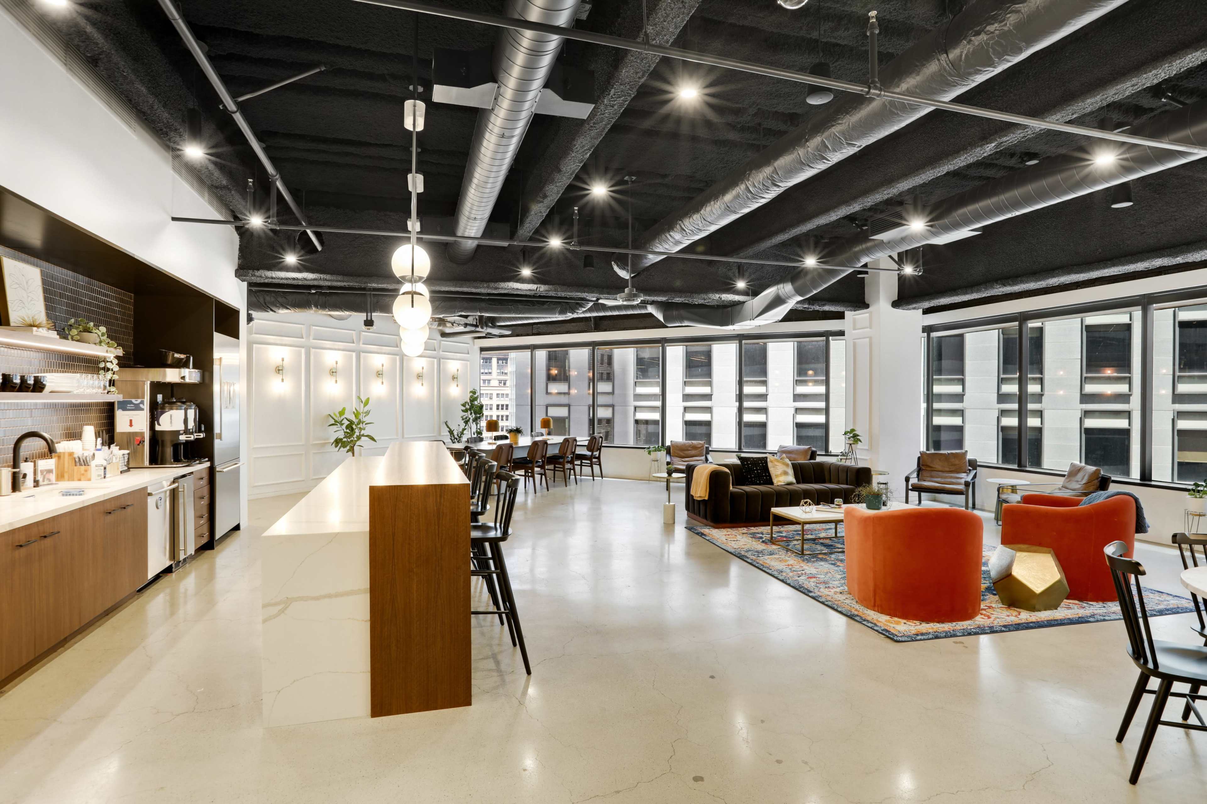 A modern workspace features a kitchen area, lounge seating, and large windows with city views.