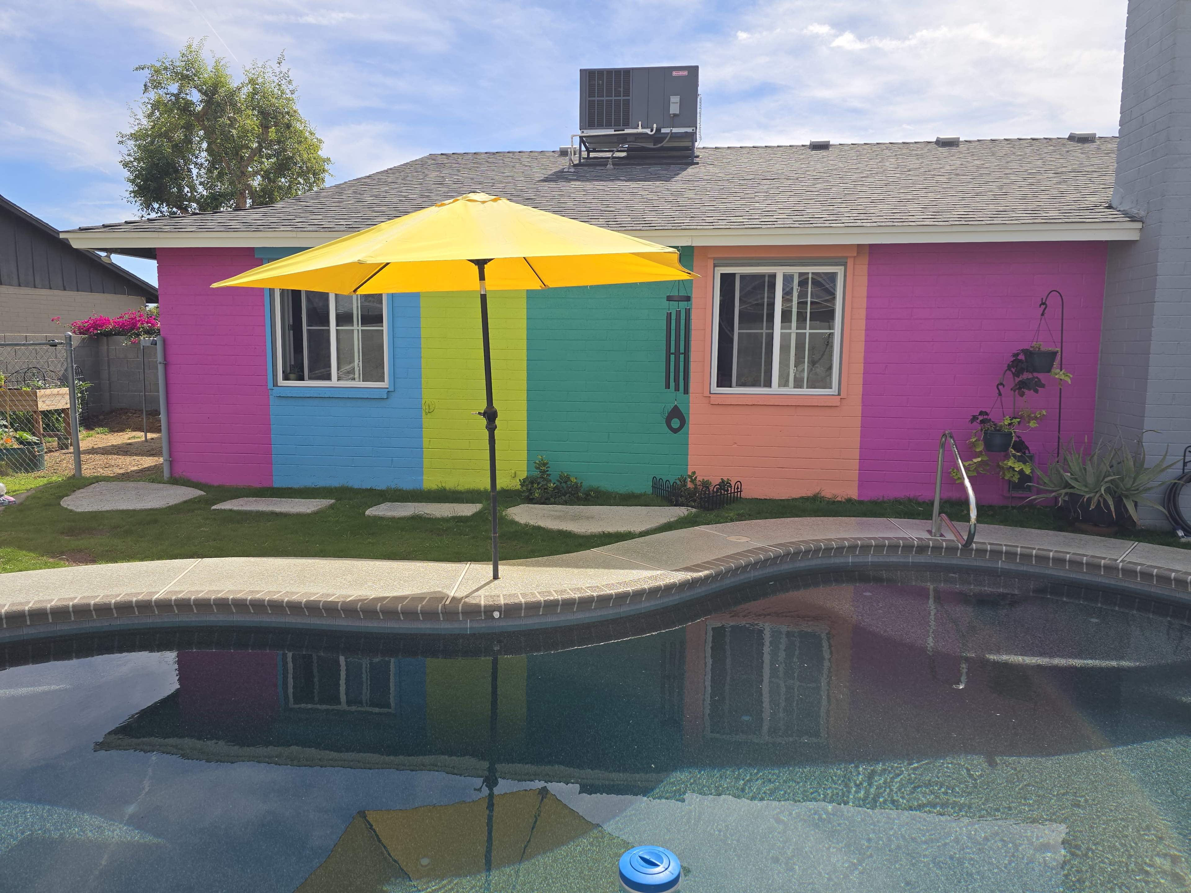 The image shows a vibrant, multicolored house with a yellow umbrella over a pool surrounded by greenery.