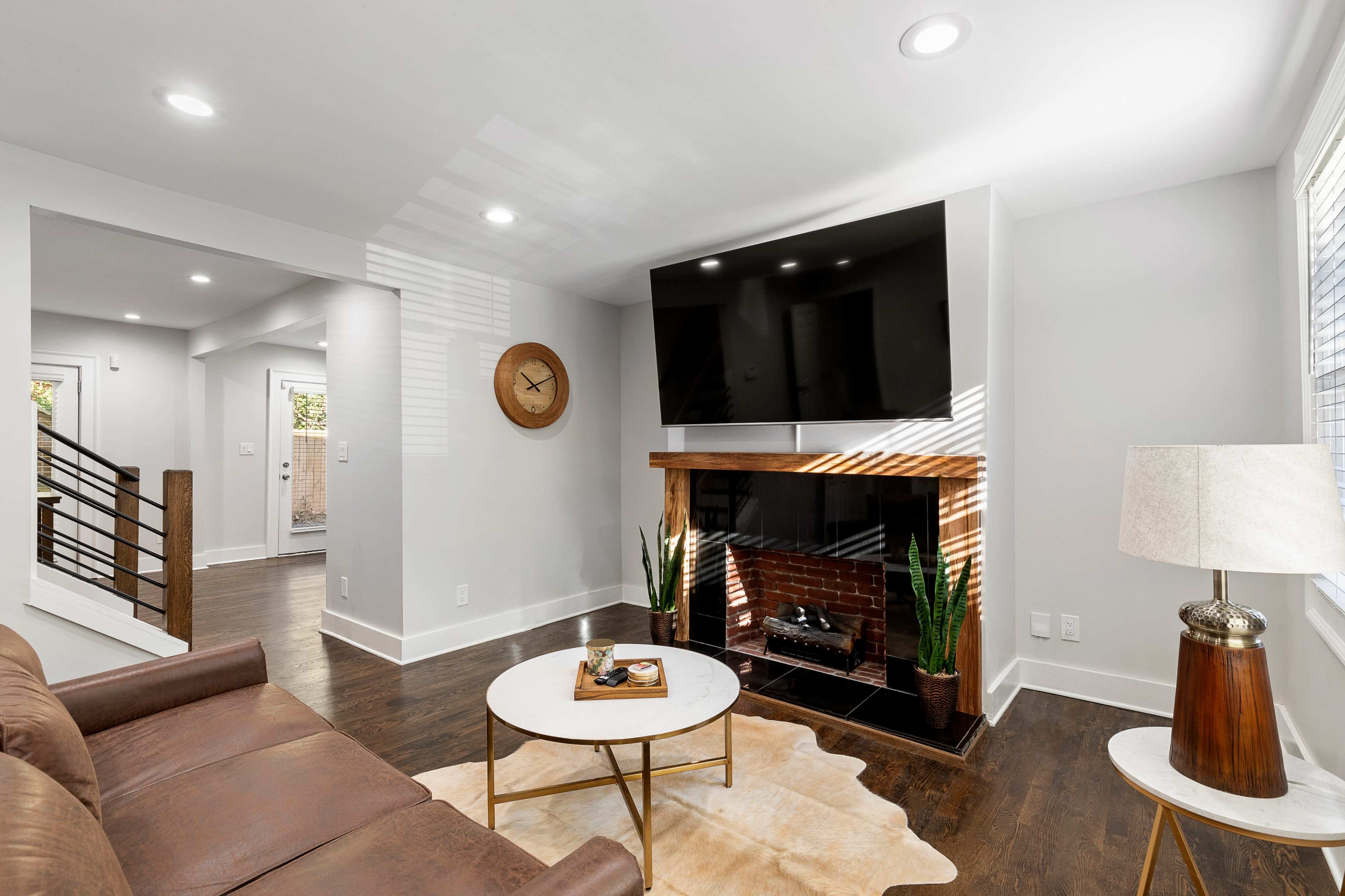 A modern living room with a brown leather sofa, a round coffee table, a wall-mounted television above a brick fireplace, and plants in the corner.