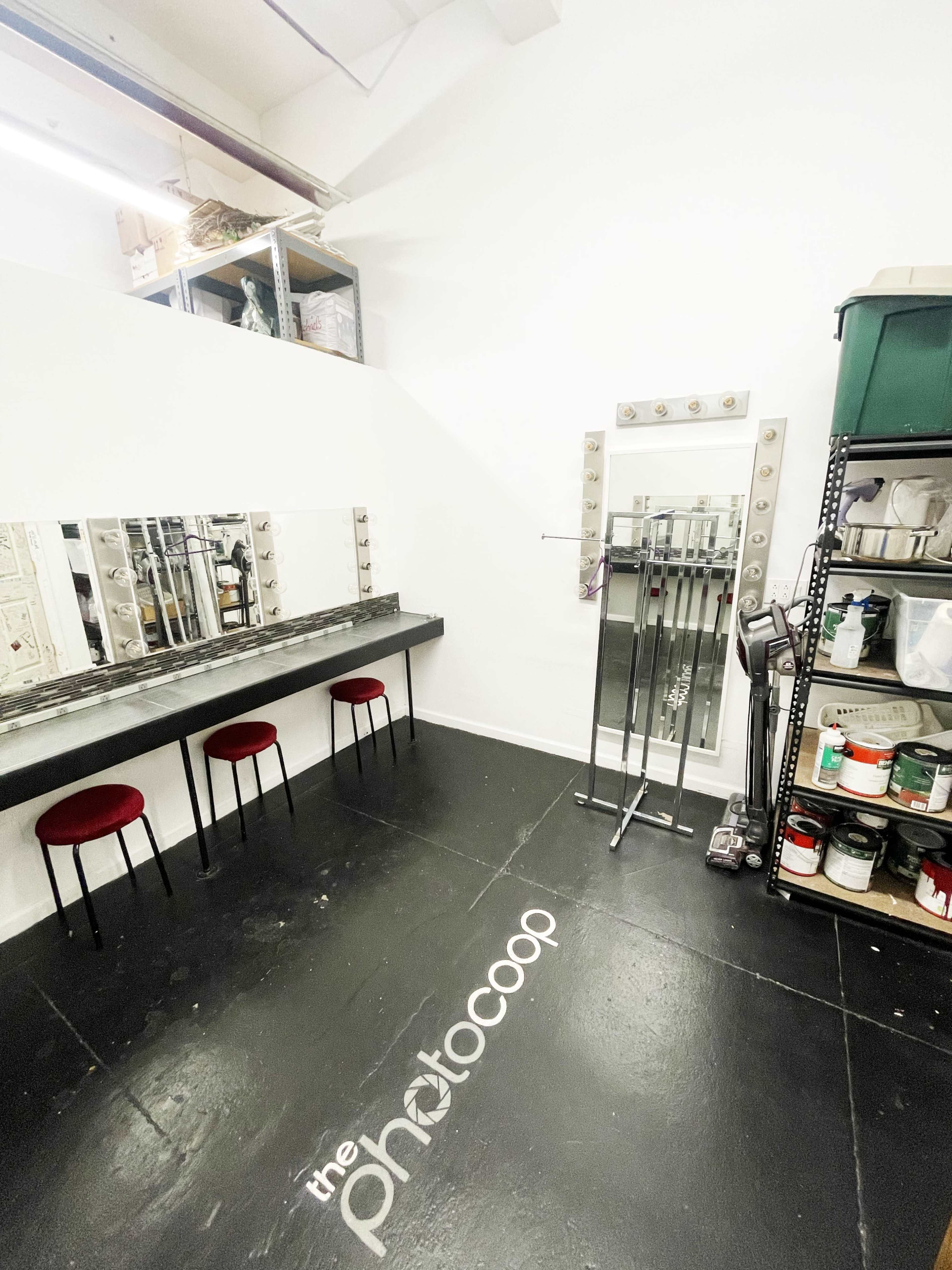 The image shows a simple dressing area with three red chairs, a large mirror with lights, and storage shelves filled with various items.