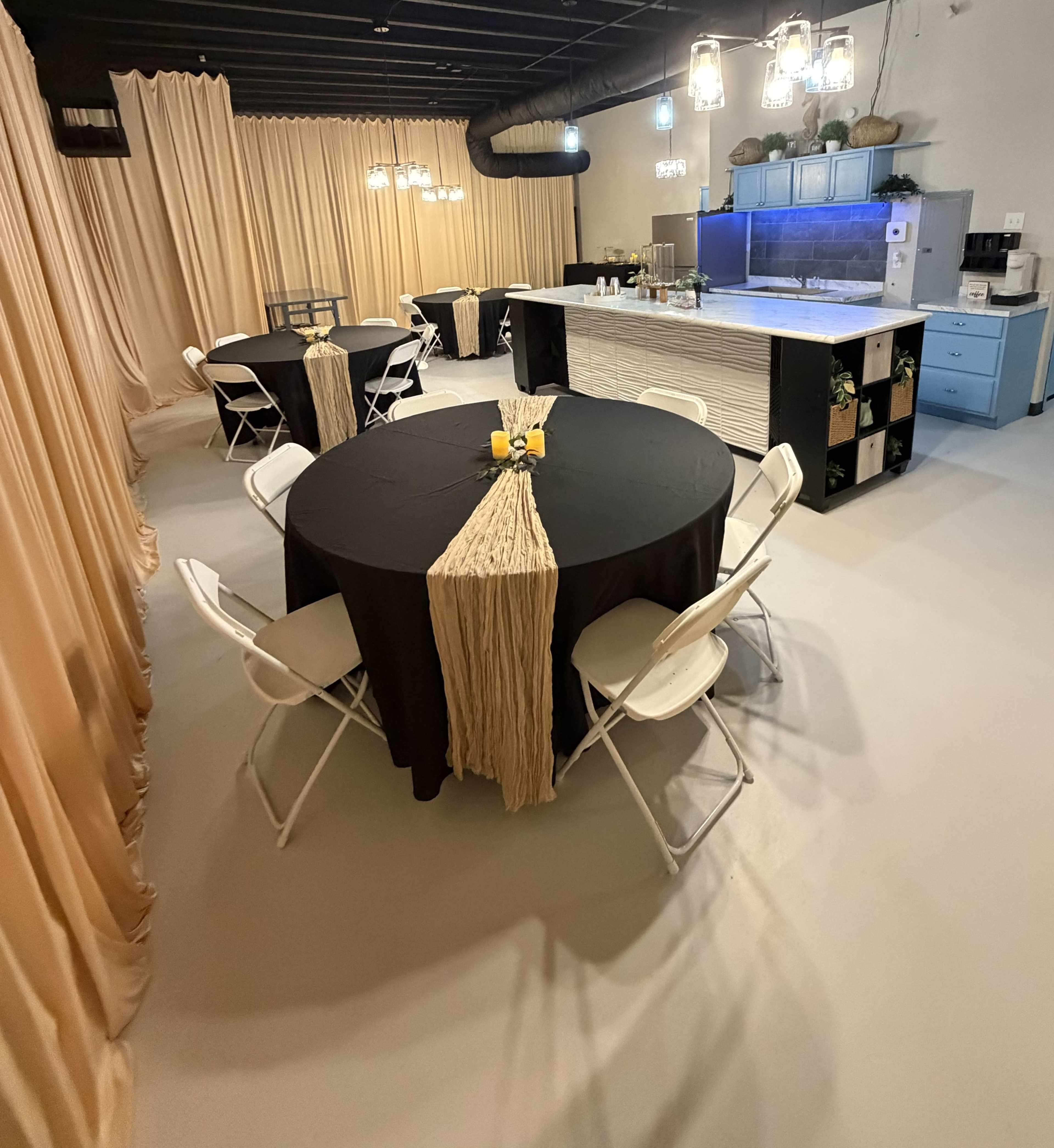 The image shows a spacious event room set up with several round black tables and white chairs, surrounded by beige curtains and featuring a kitchen area with a blue cabinetry.