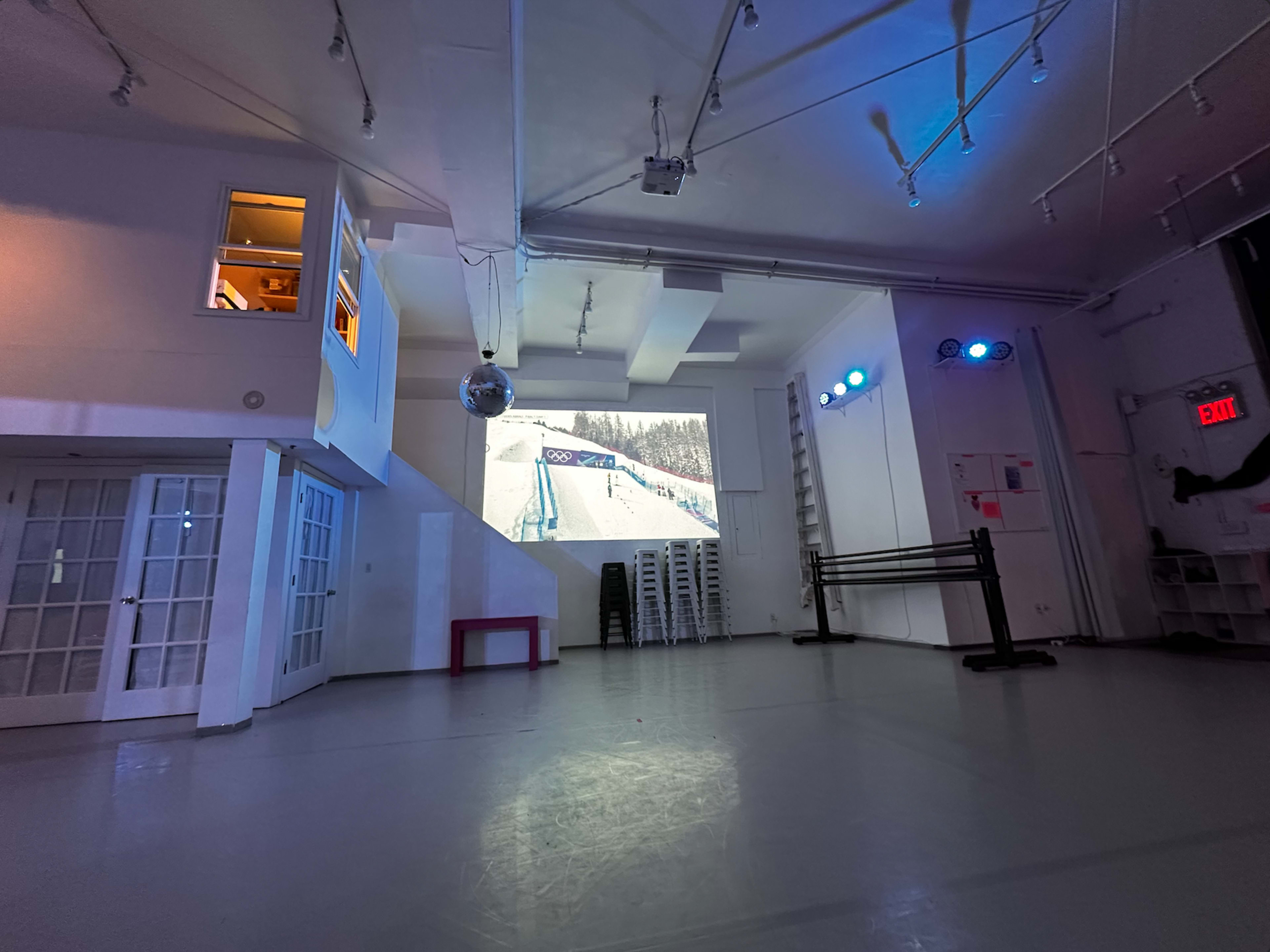 The image shows an empty dance studio with a large projection screen displaying a snowy mountain scene, illuminated by blue lights and featuring a disco ball hanging from the ceiling.