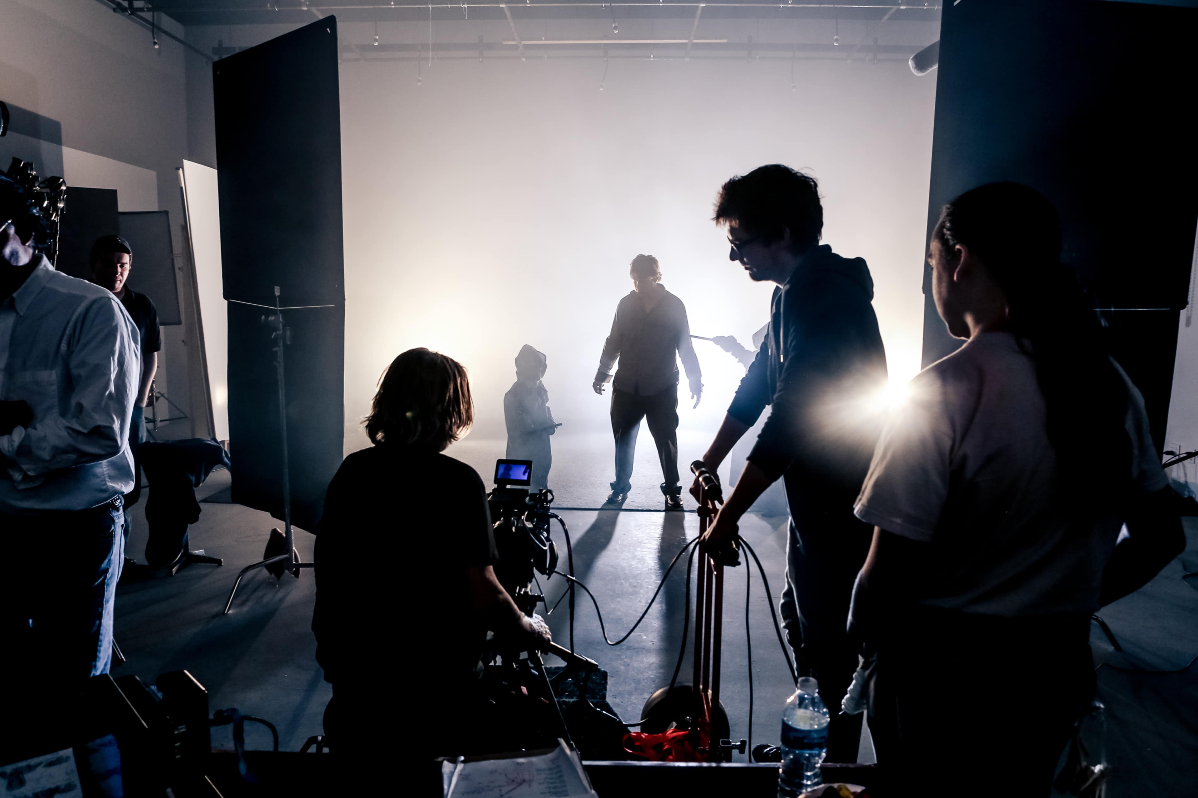 A film crew works in a dimly lit studio, with silhouetted figures and equipment set up for a shoot.