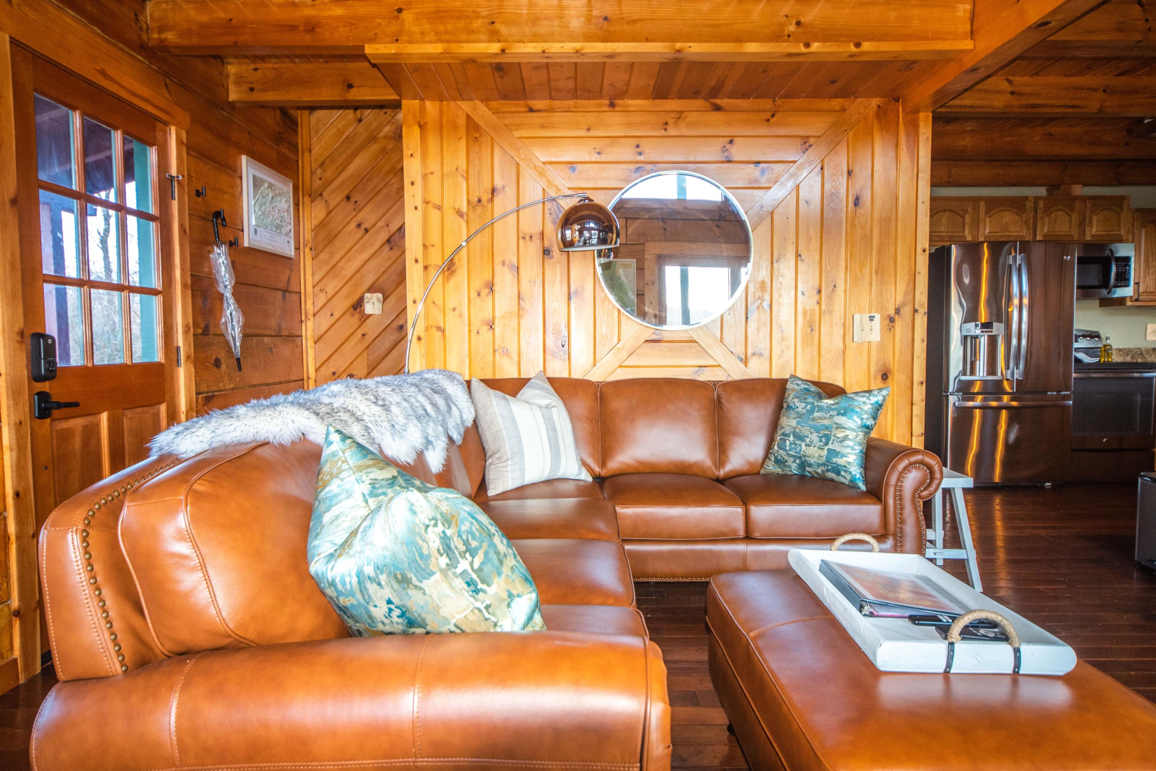 A brown leather sectional sofa with decorative pillows is situated in a wooden-paneled living room featuring a round mirror and a coffee table.
