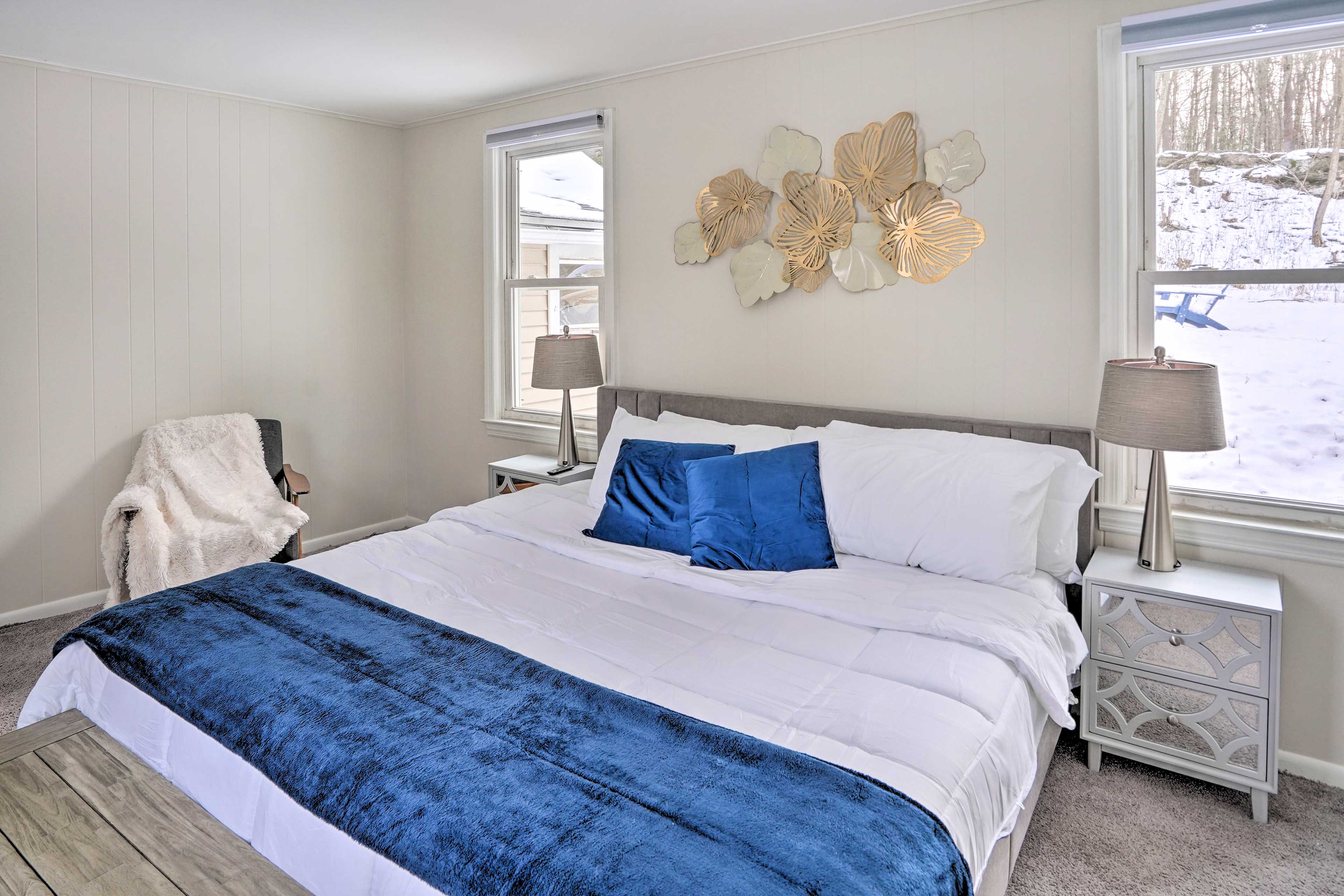 A bedroom features a queen-sized bed with white linens and a blue throw blanket, flanked by lamps and a decorative wall piece, with windows overlooking a snowy landscape.
