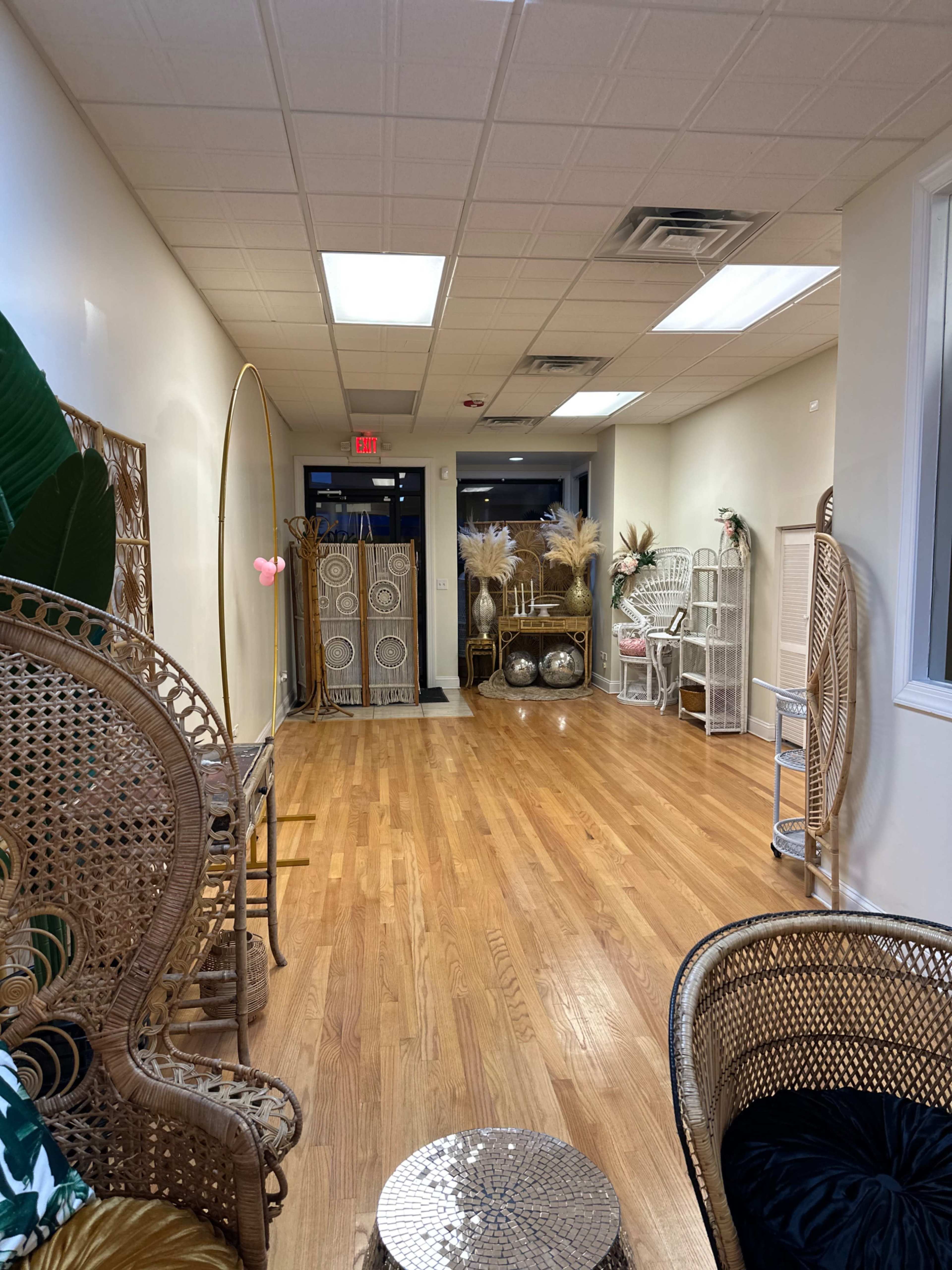 The image shows a well-lit hallway featuring various decorative wicker furniture and decor arranged along the walls, with wooden floors and large windows at the far end.