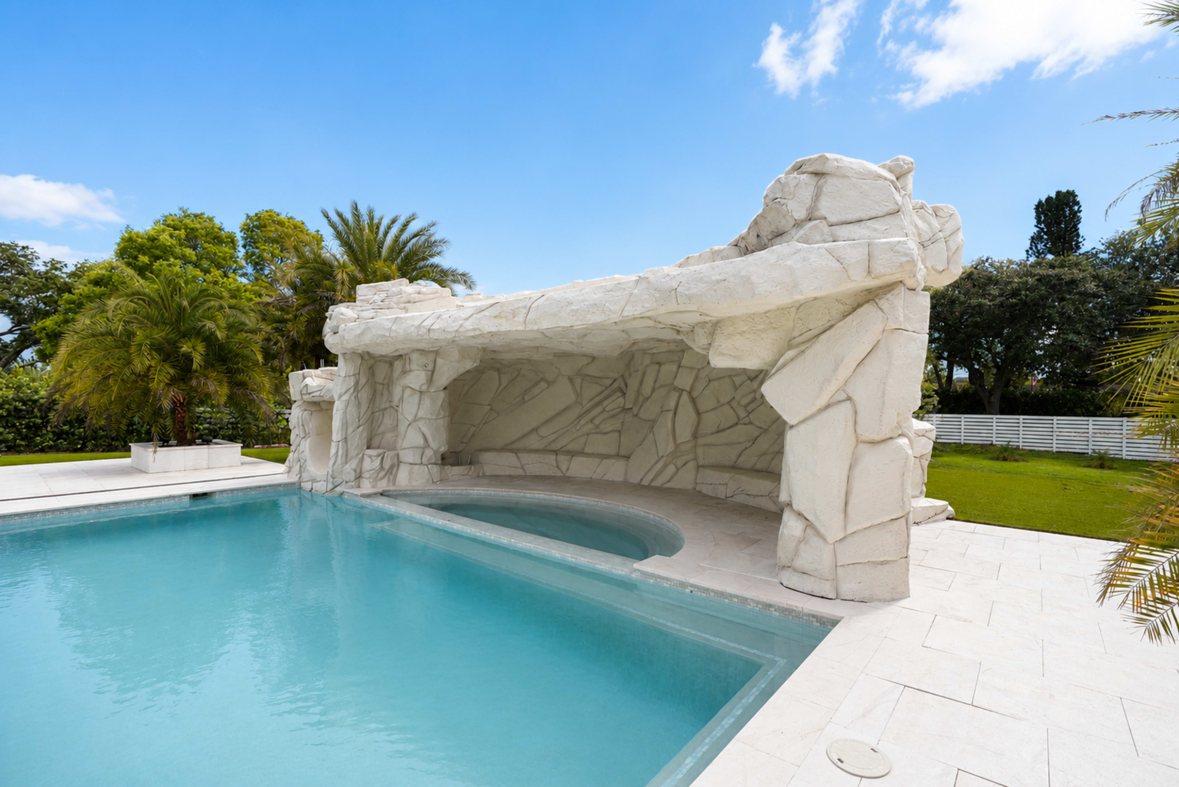 The image shows a swimming pool with a white stone cave-like structure and palm trees in the background under a clear blue sky.