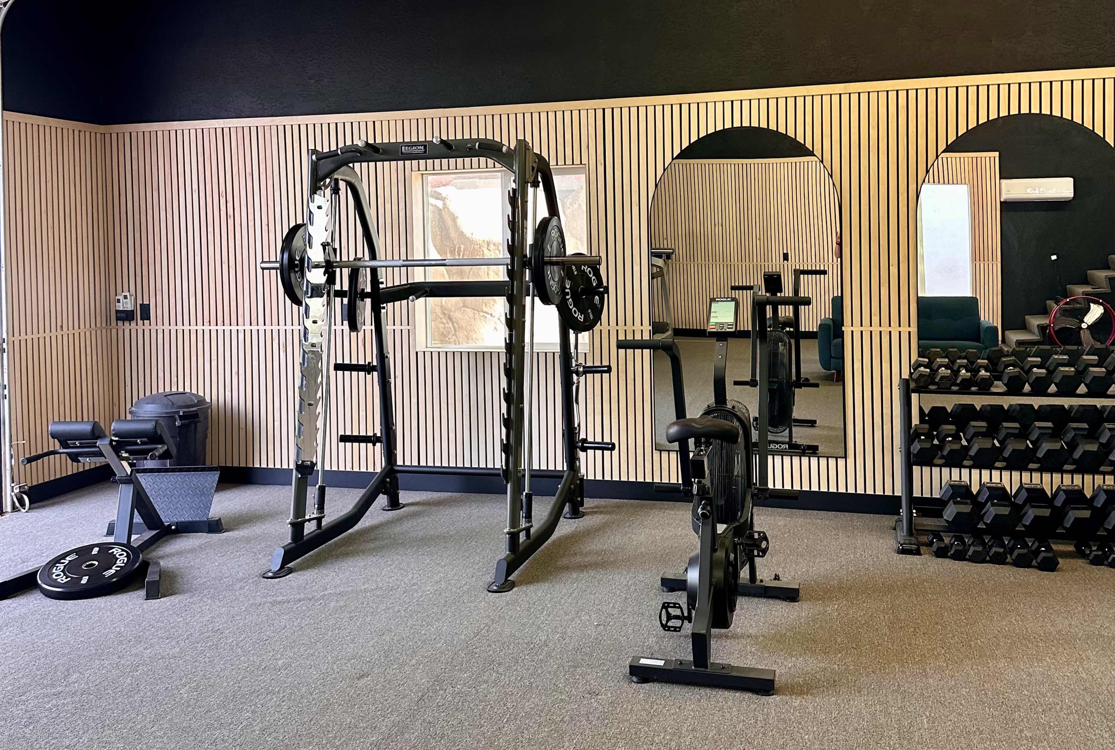 The image shows a gym interior featuring a squat rack, a weight bench, a set of dumbbells on a rack, and mirrors along the wall.