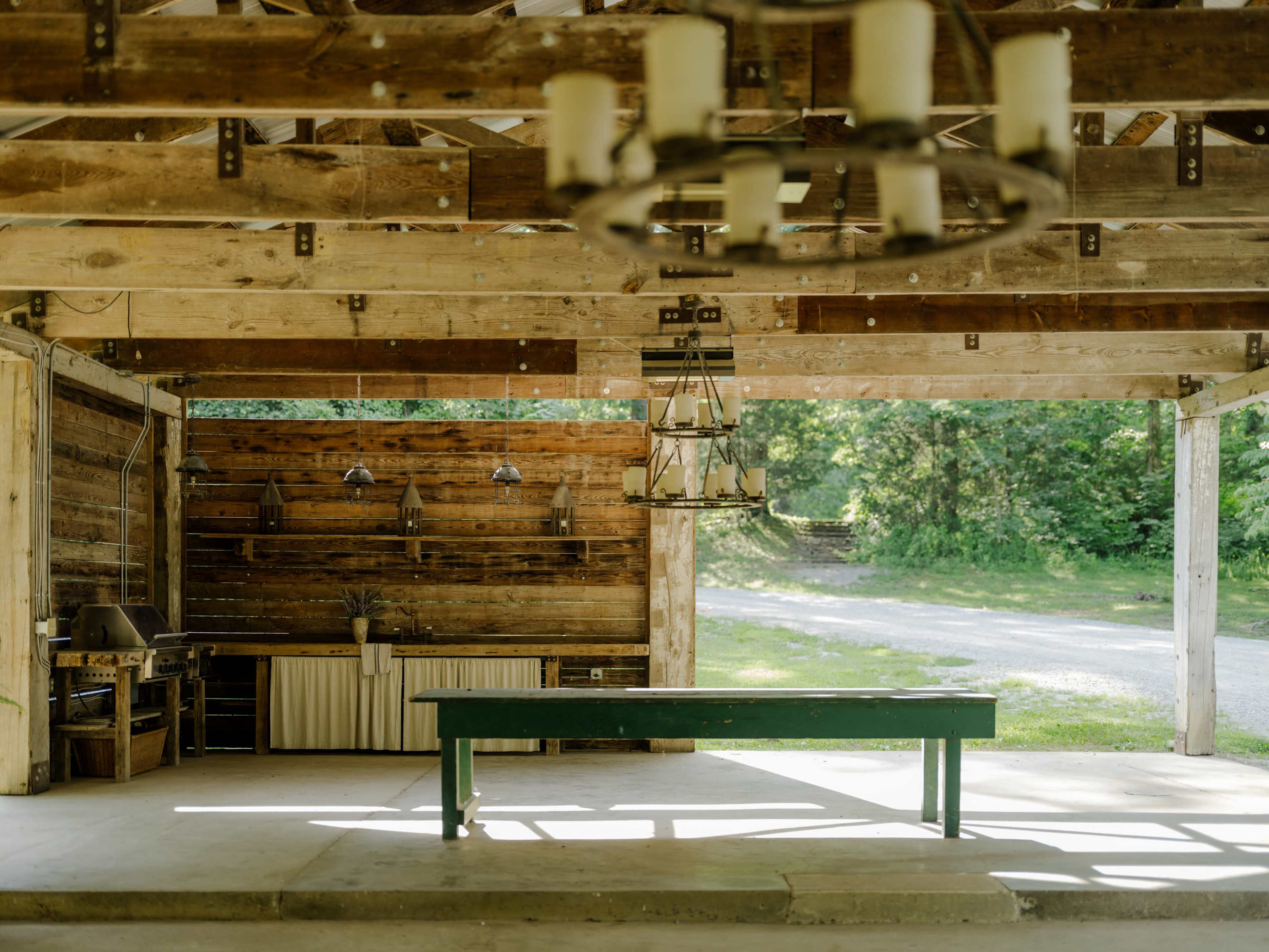 The image shows a spacious, rustic outdoor pavilion with wooden beams, a large work table, and a kitchenette area against a backdrop of greenery.