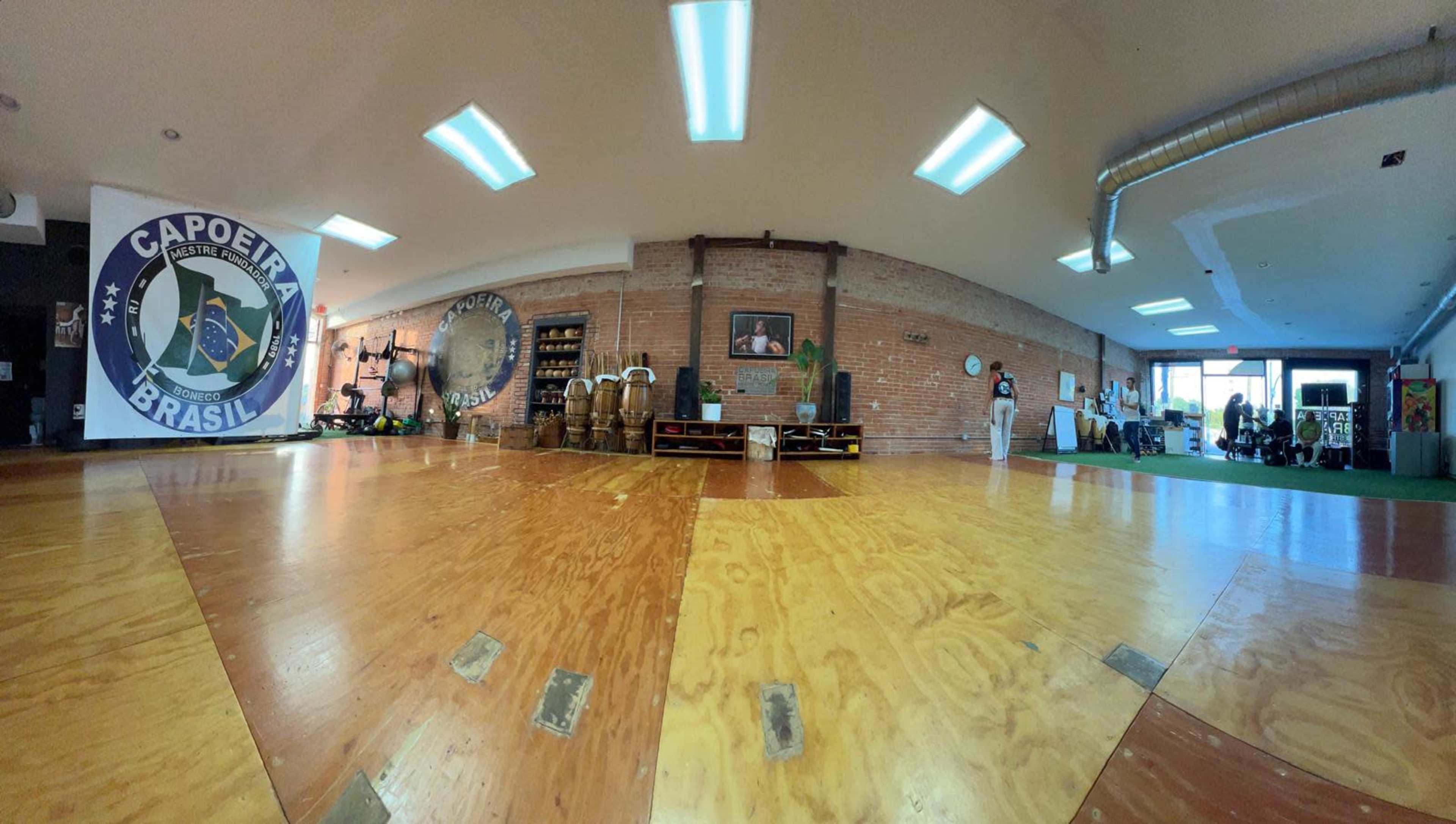 The image shows a spacious room designed for Capoeira practice, featuring wooden flooring, wall murals, and various training equipment in an organized setting.
