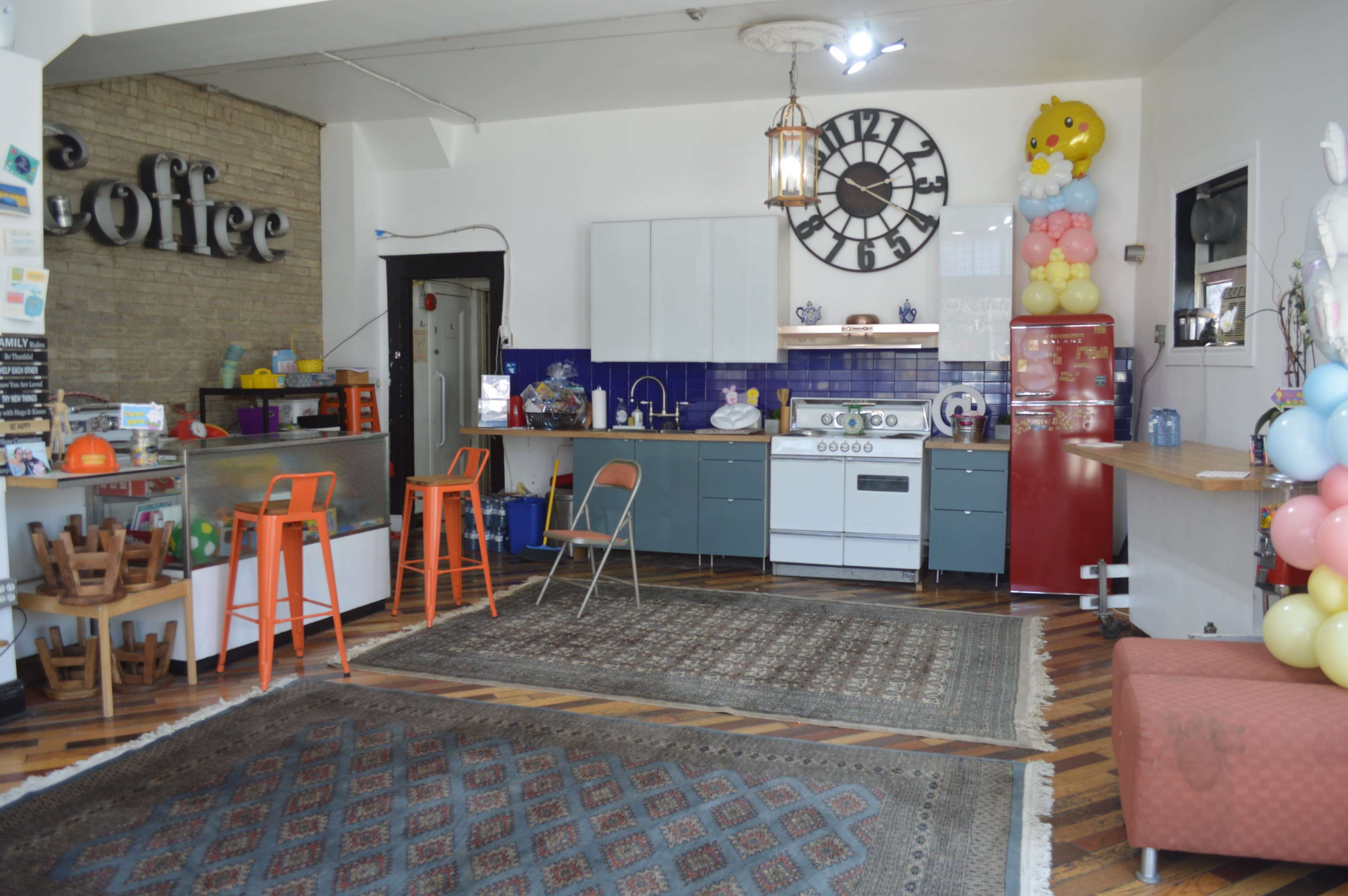 A café interior with a modern kitchen area, featuring a blue and gray color scheme, a vintage clock on the wall, and colorful decorations, including balloons.