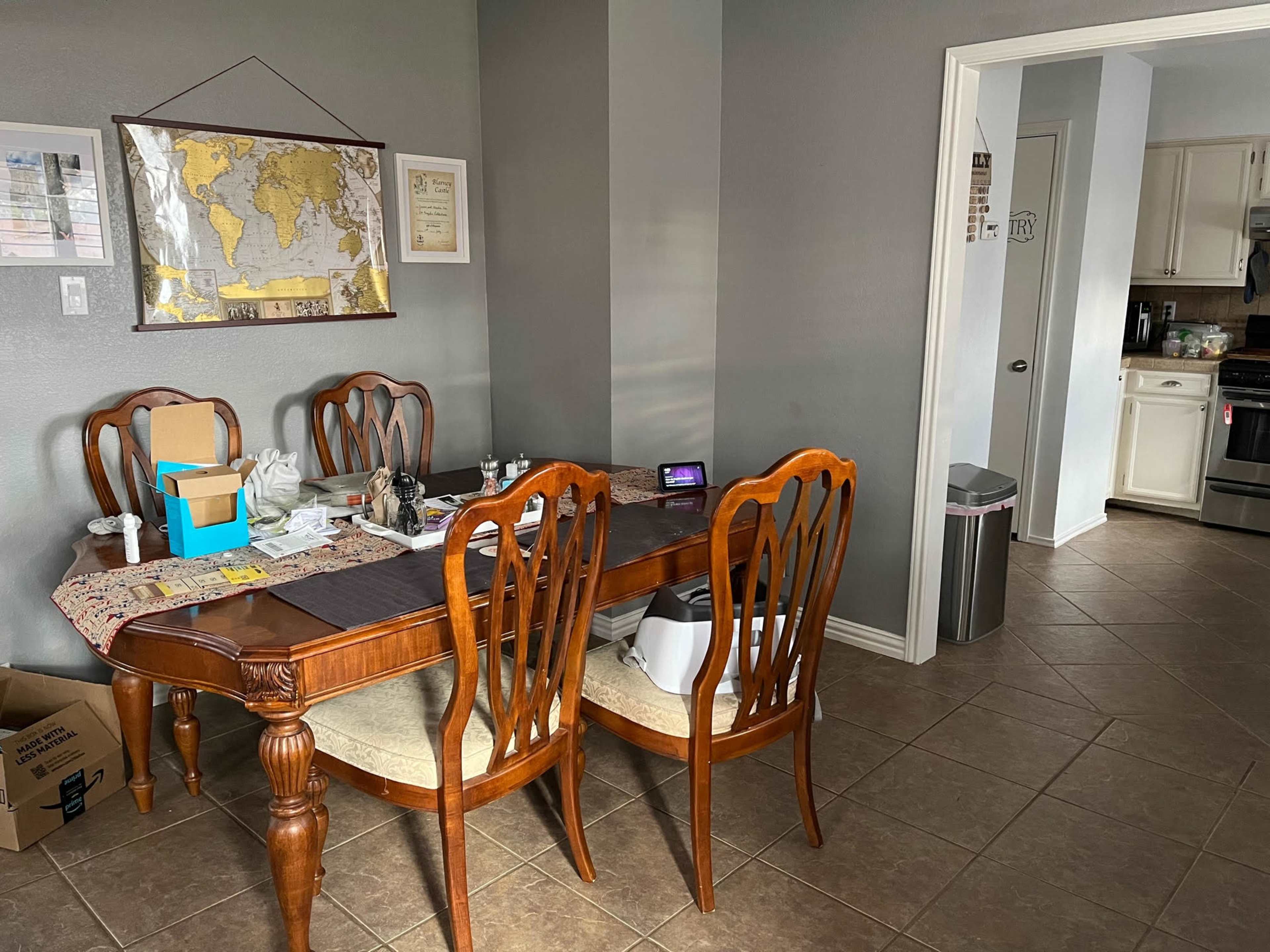 A wooden dining table with chairs is positioned in a well-lit room, featuring a map on the wall and a doorway leading to a kitchen.