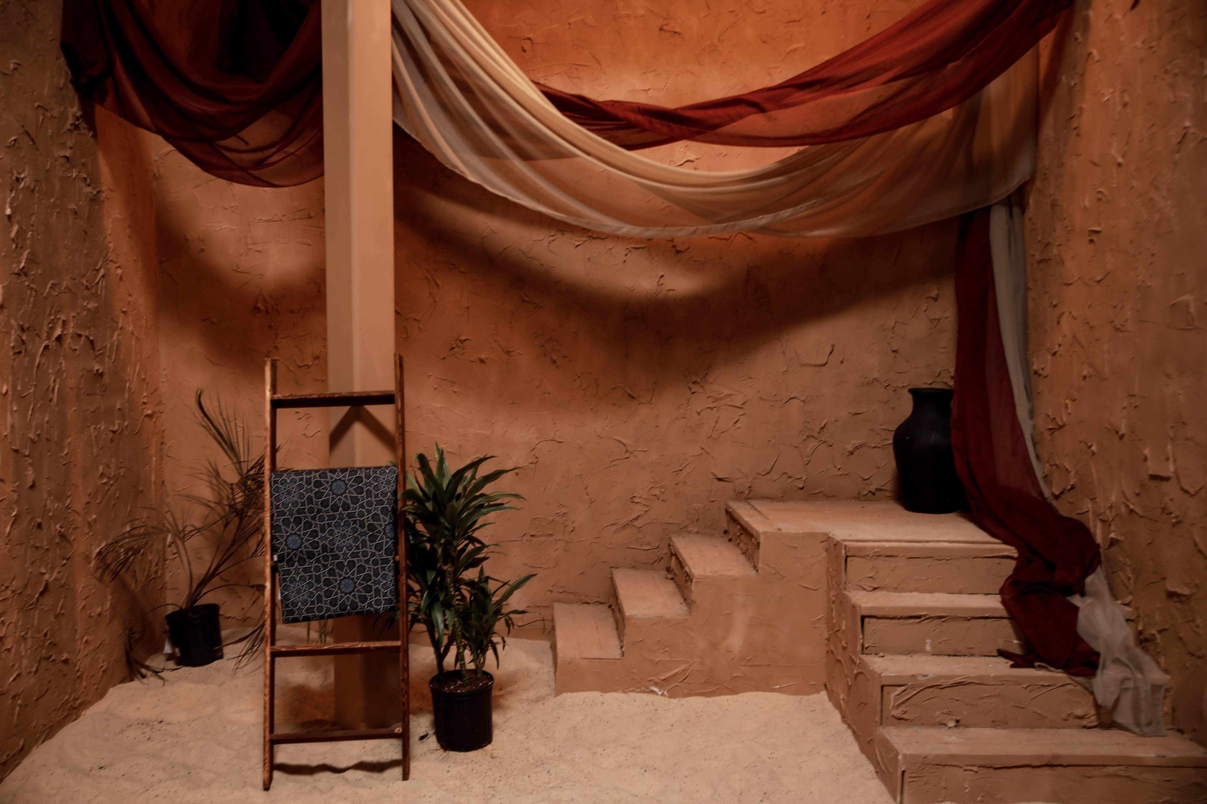 An interior setting with textured walls in warm colors, a staircase, a folding chair, a potted plant, and draped fabric hanging from the ceiling.