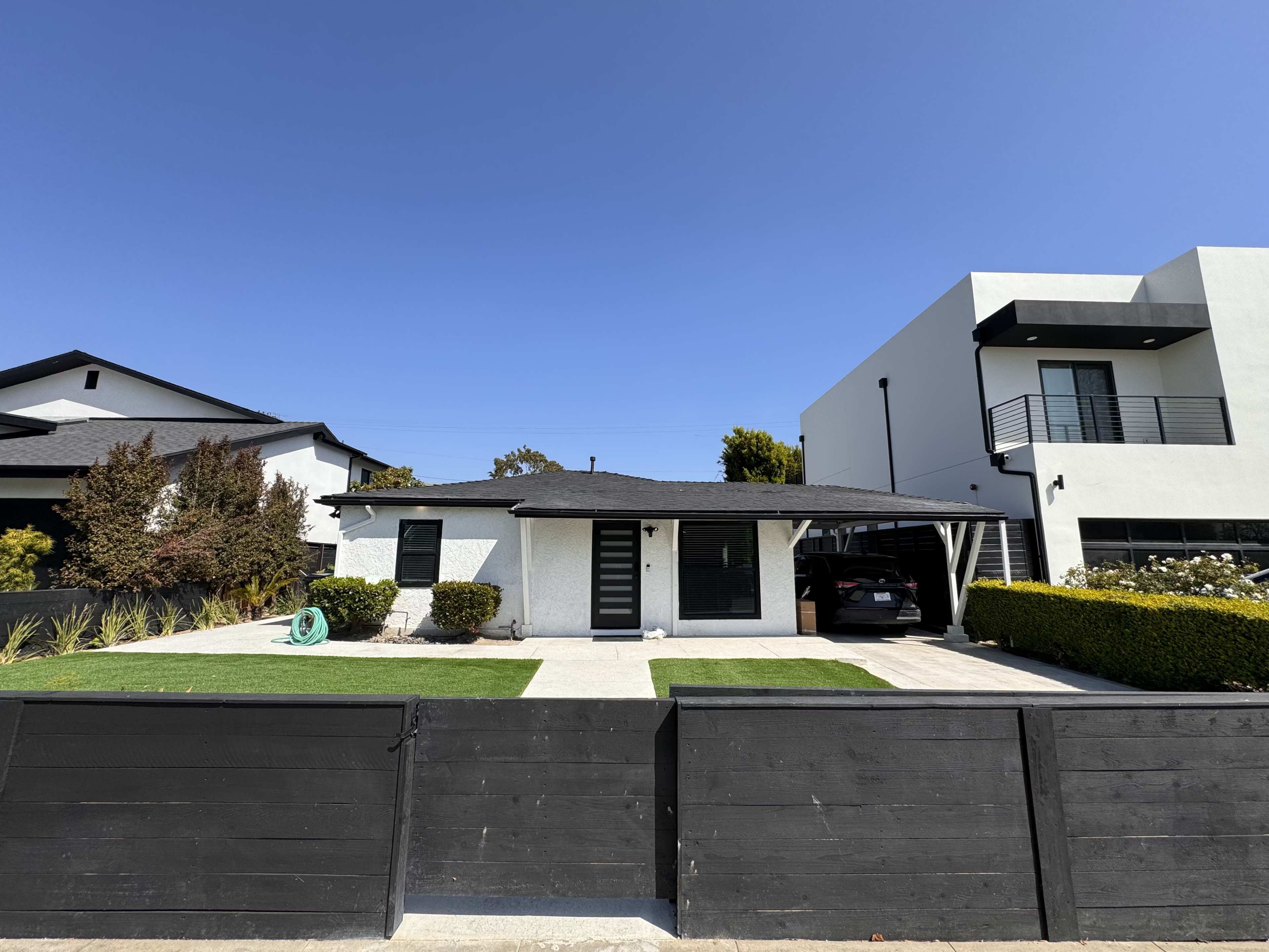 The image shows a single-story white house with a black roof, situated between two modern buildings, with a manicured lawn and a black fence in front.