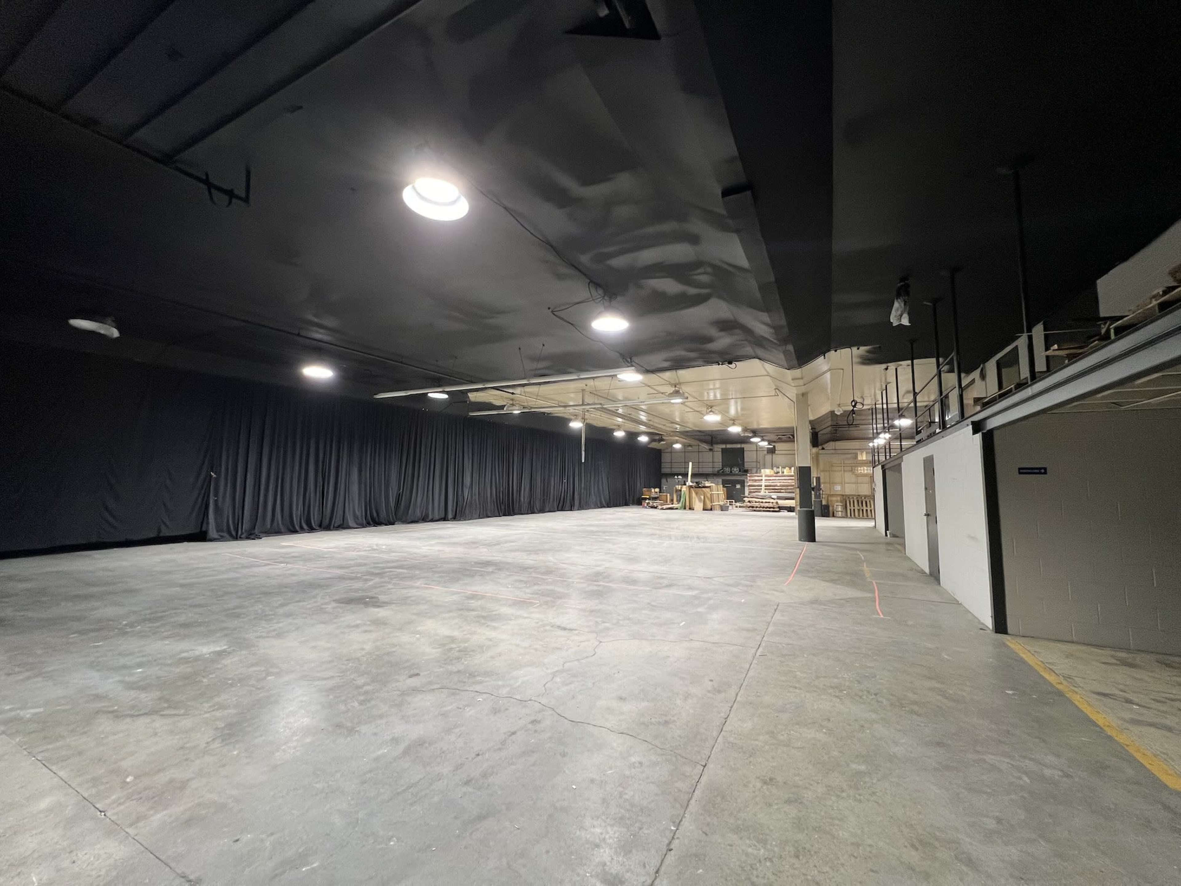 The image shows a large, empty warehouse space with black curtains along one wall and concrete floors.