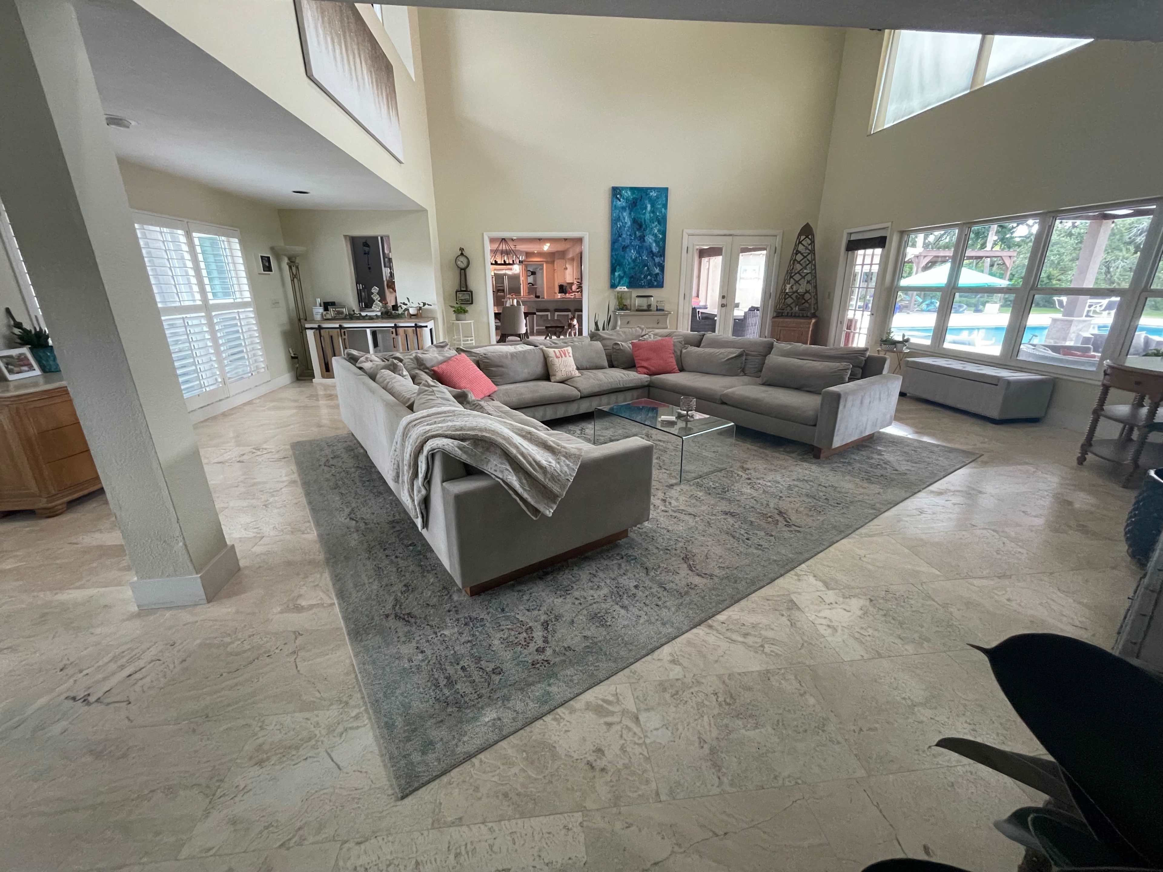 A spacious living room features a large sectional sofa arranged around a glass coffee table, with large windows allowing natural light and a view of an outdoor area.
