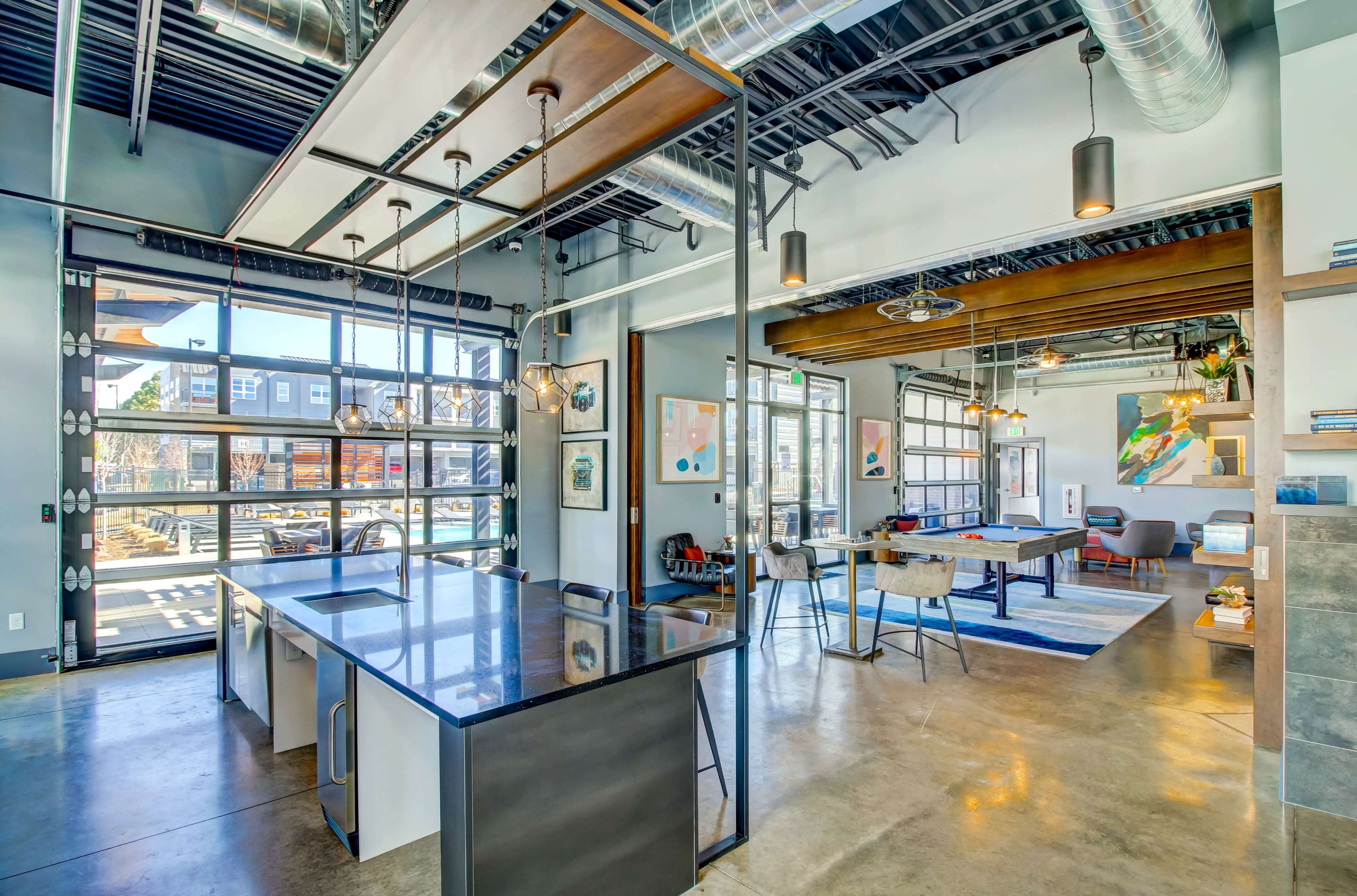A modern open-concept office space with large windows, industrial lighting, and a kitchen area adjacent to collaborative workspaces.