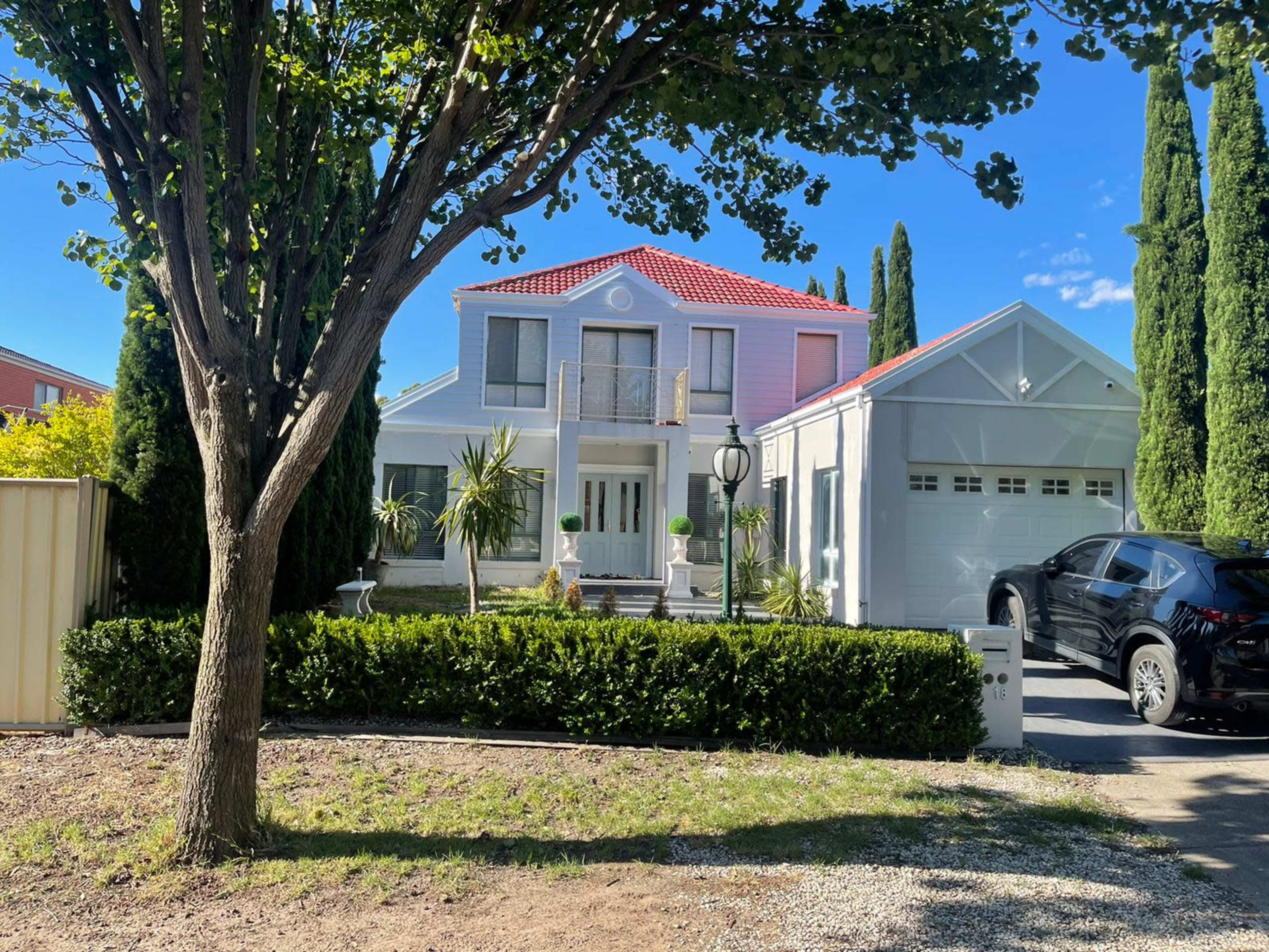 A two-story house with a red-tiled roof is surrounded by tall trees and shrubs, with a garage on the right and a car parked in the driveway.